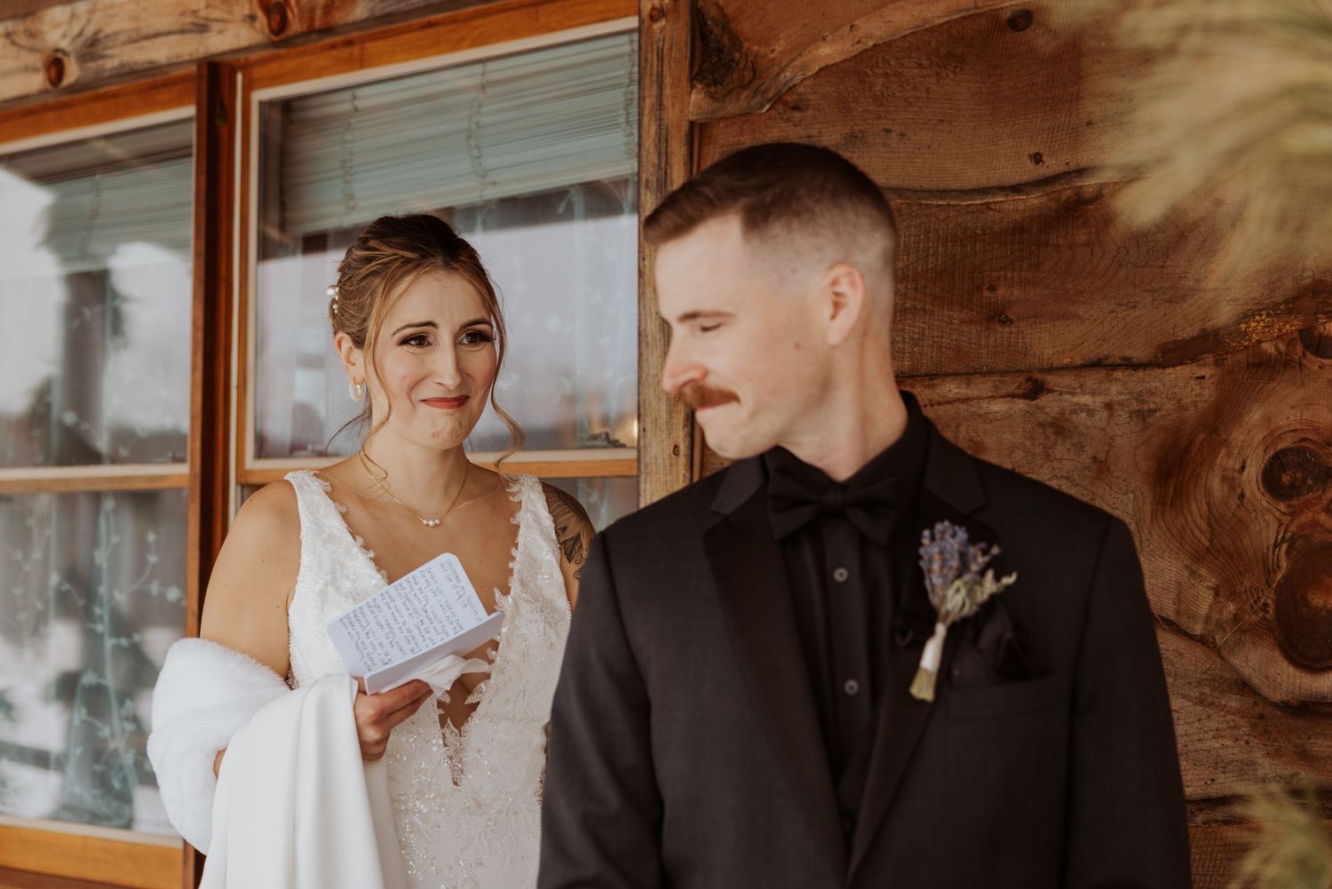 Bride reading vows to groom outside rustic log cabin wedding venue, groom wearing black suit with floral boutonniere.