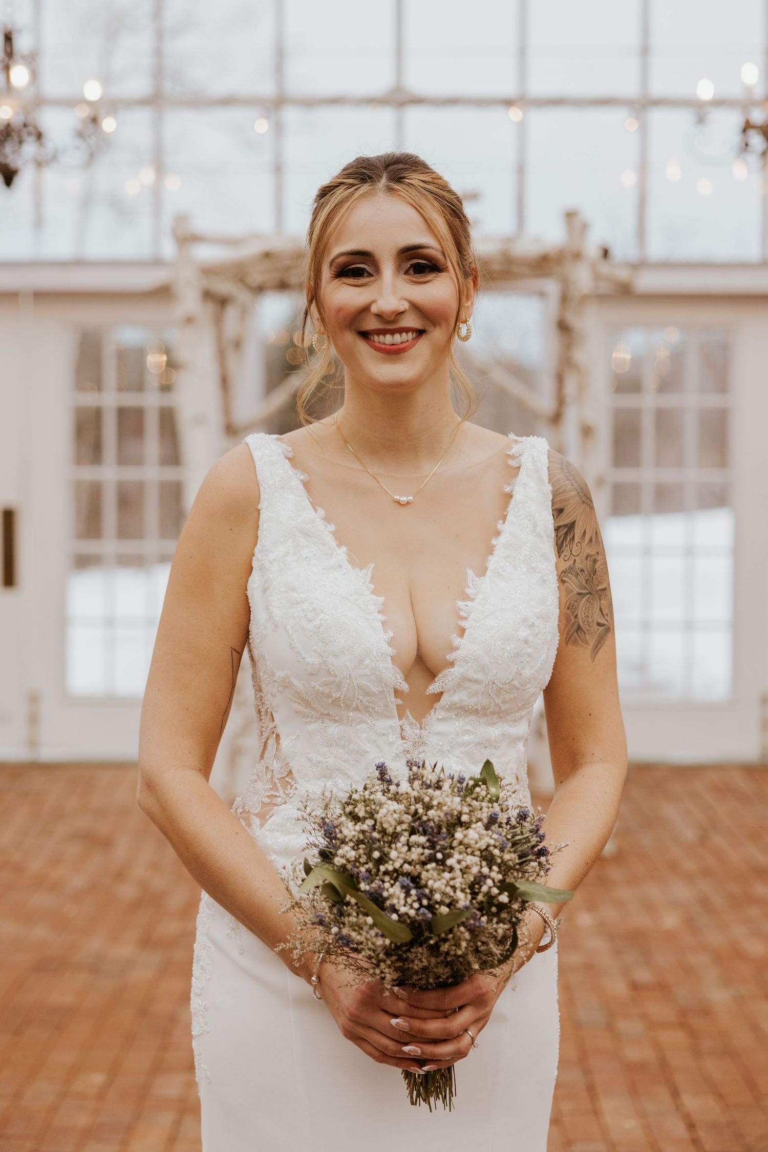 Smiling bride in lace wedding gown holding wildflower bouquet inside a greenhouse venue.