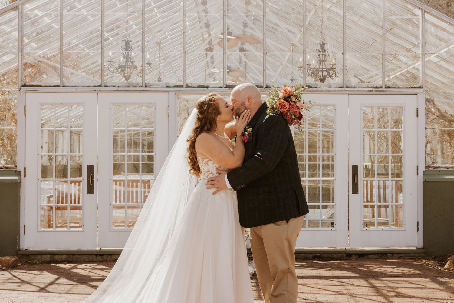A couple shares an intimate moment in front of a sunlit greenhouse conservatory with elegant white architecture.