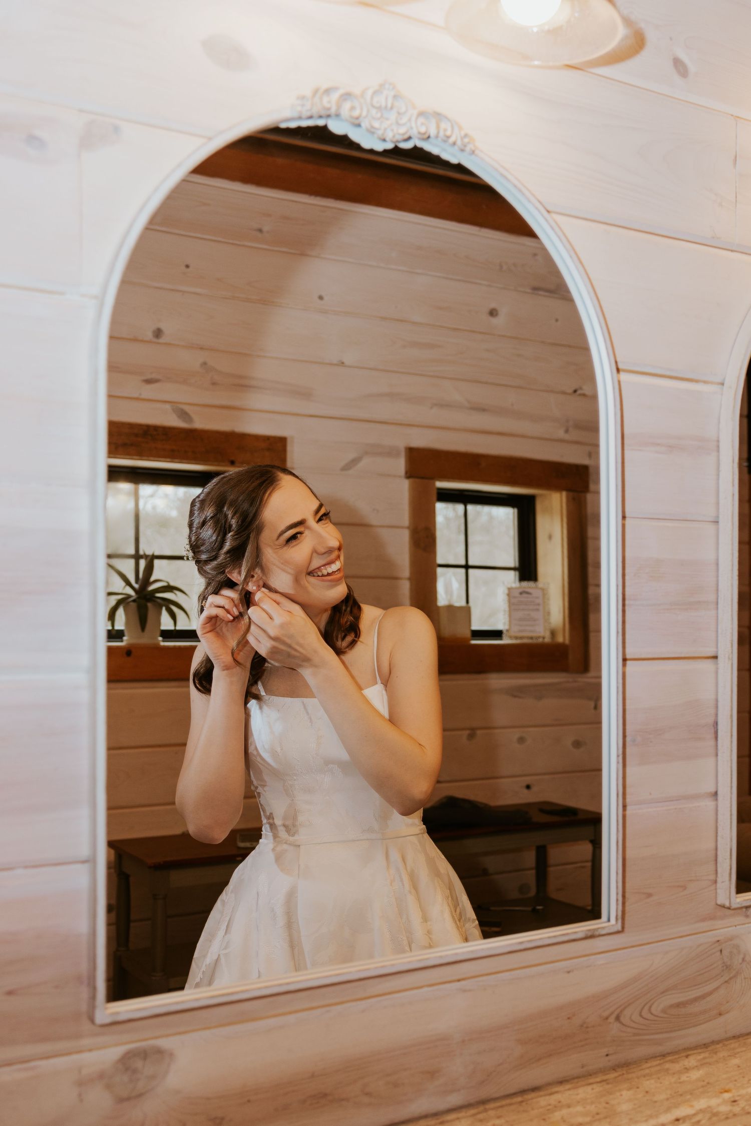 A bride in a white dress smiles while getting ready in a rustic wooden room with an arched window frame.
