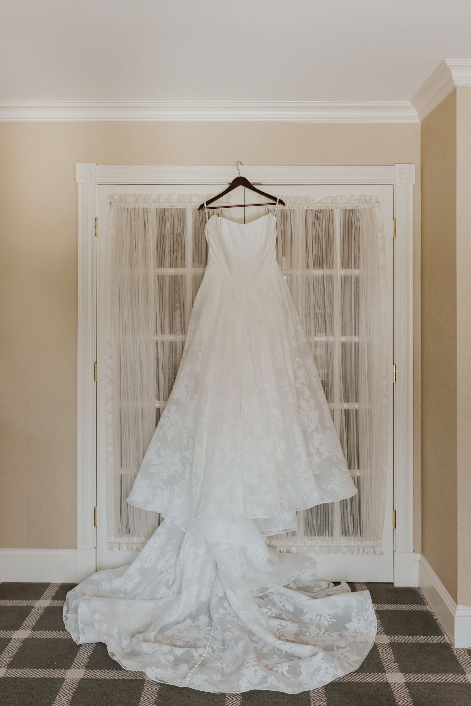 A strapless lace wedding gown with a long train hangs elegantly on a door in a softly lit room.
