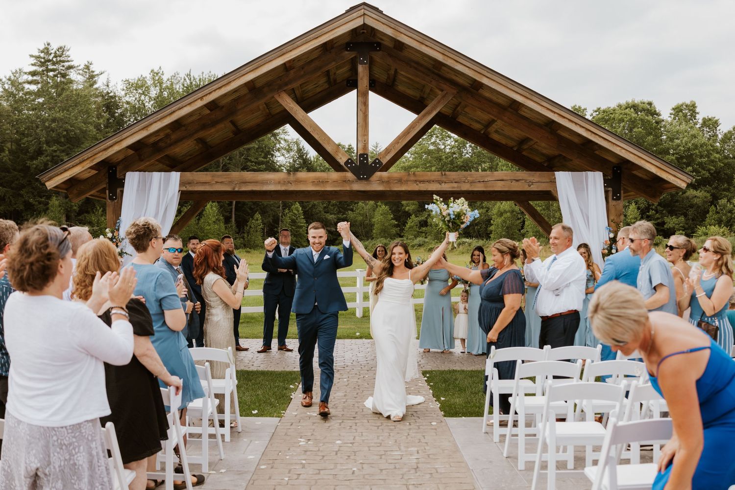 Joyful newlyweds raise hands as they exit their outdoor ceremony under a rustic wooden pavilion.