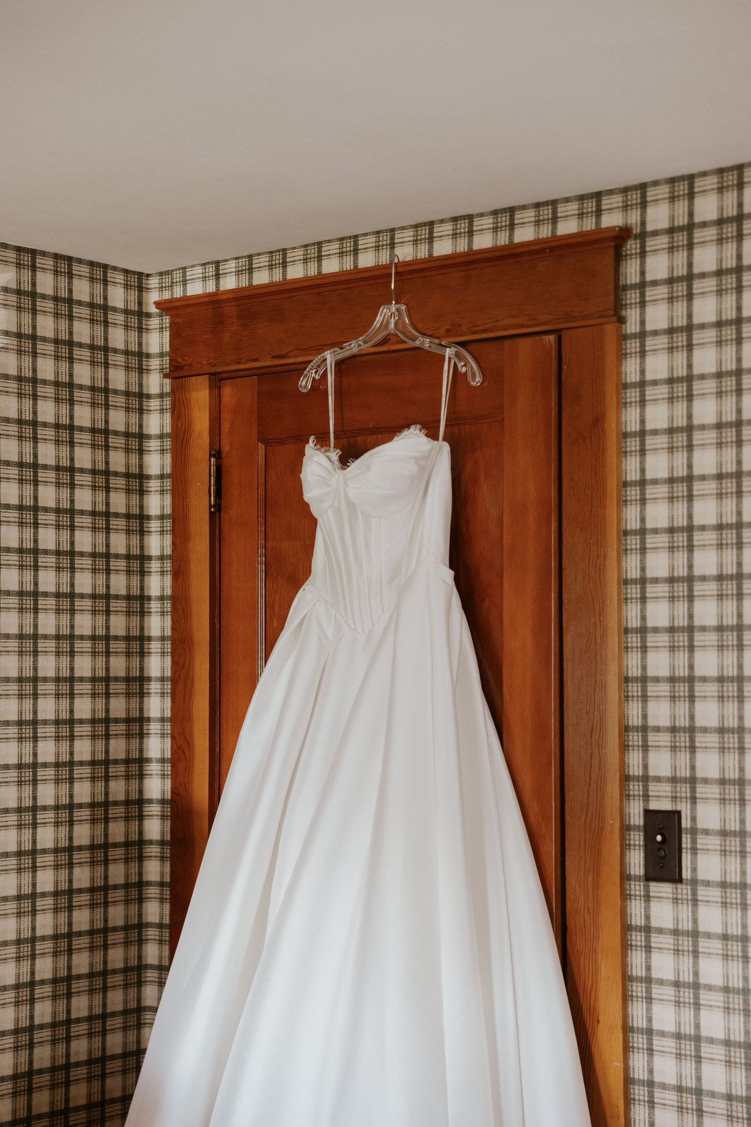 A strapless white wedding ball gown hanging on a wooden door against plaid wallpaper.