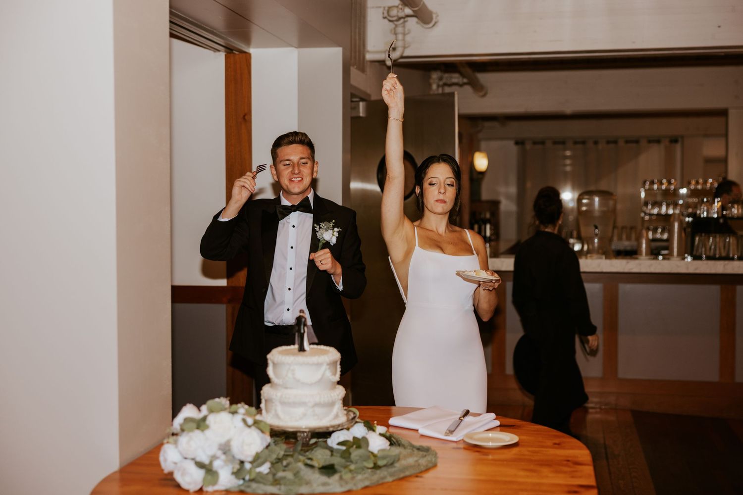 Bride and groom posing with white tiered wedding cake at reception.