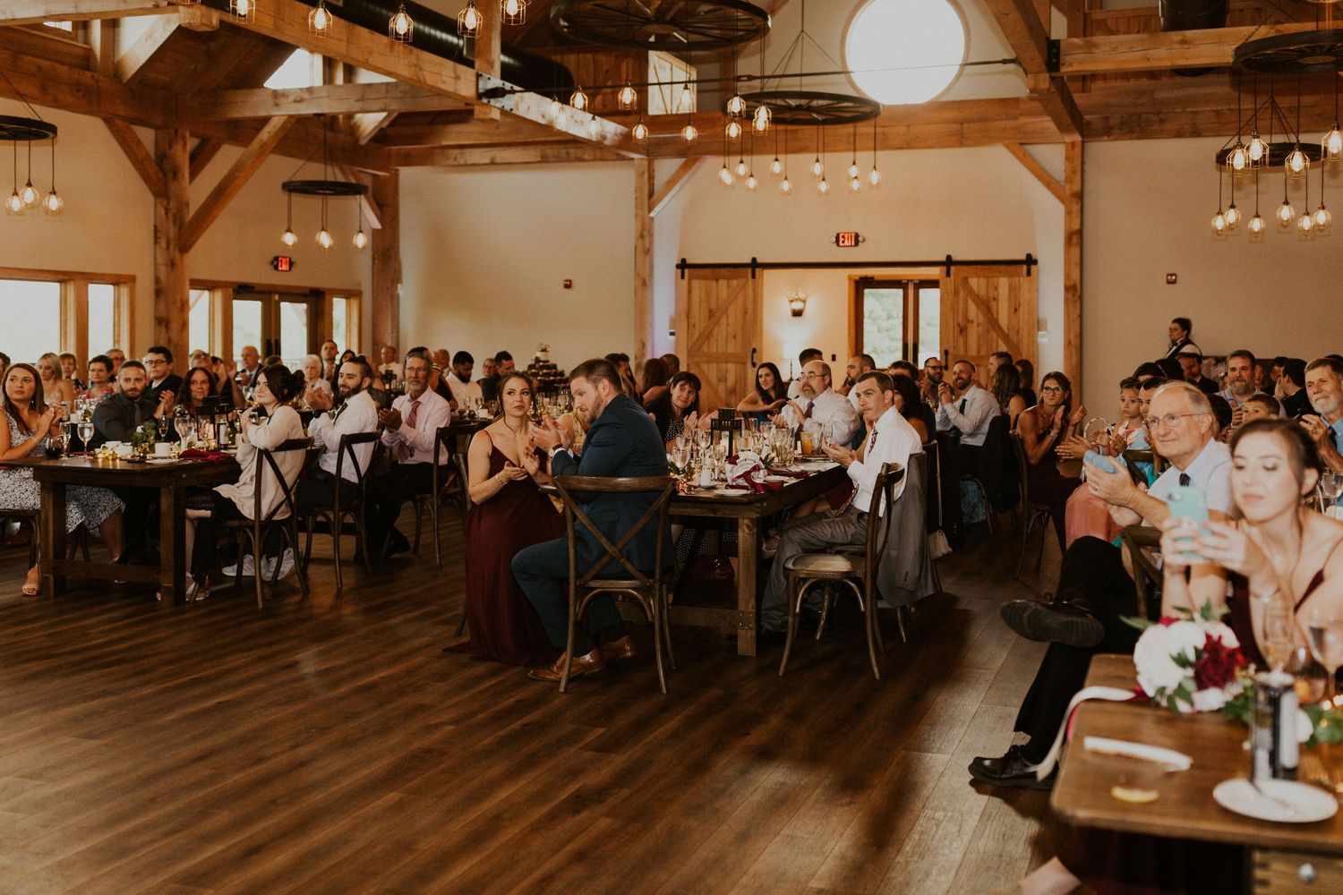 Large reception hall filled with wedding guests seated at round tables under wooden beams.