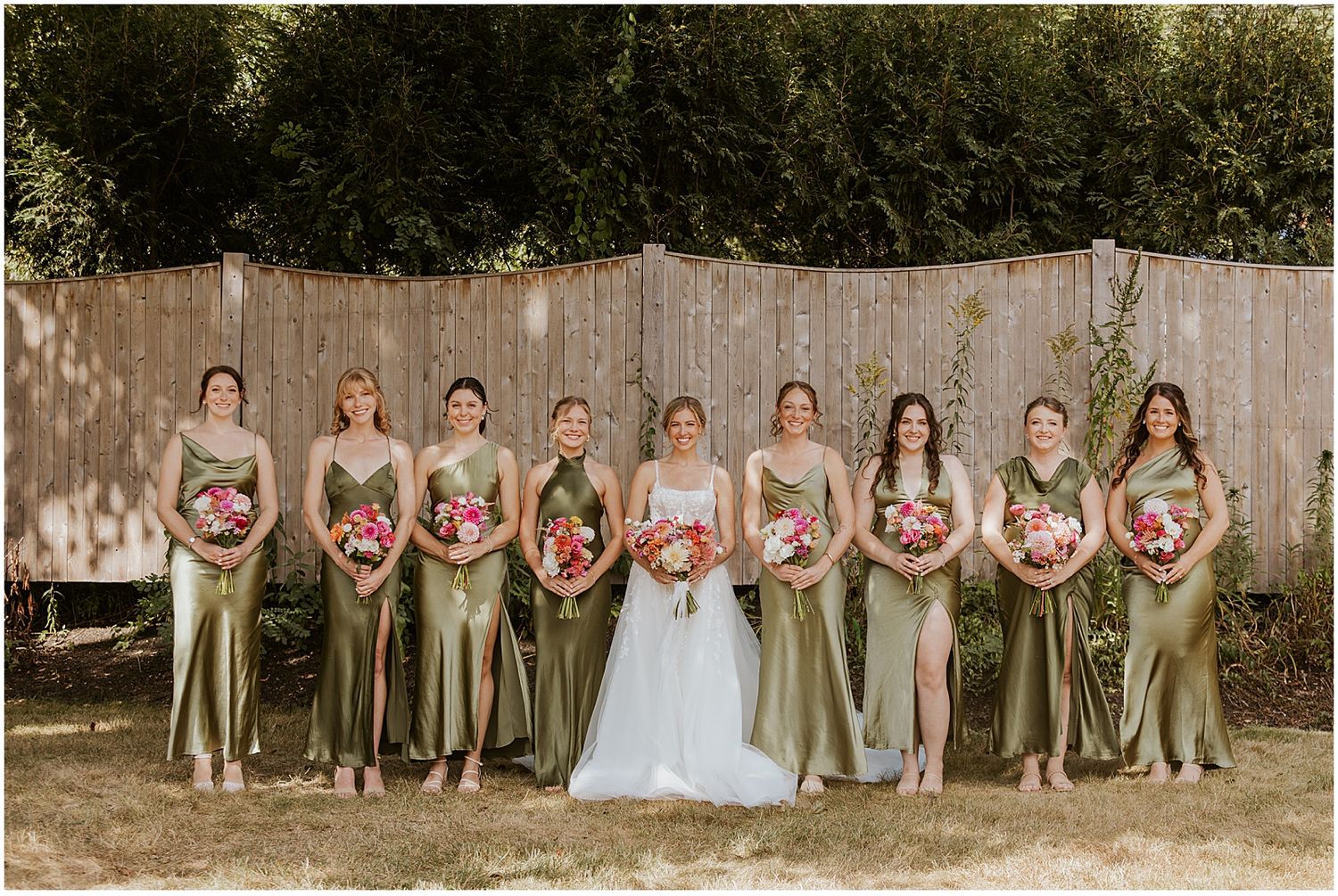 Wedding party in sage green dresses and light suits pose together against a rustic wooden fence backdrop.