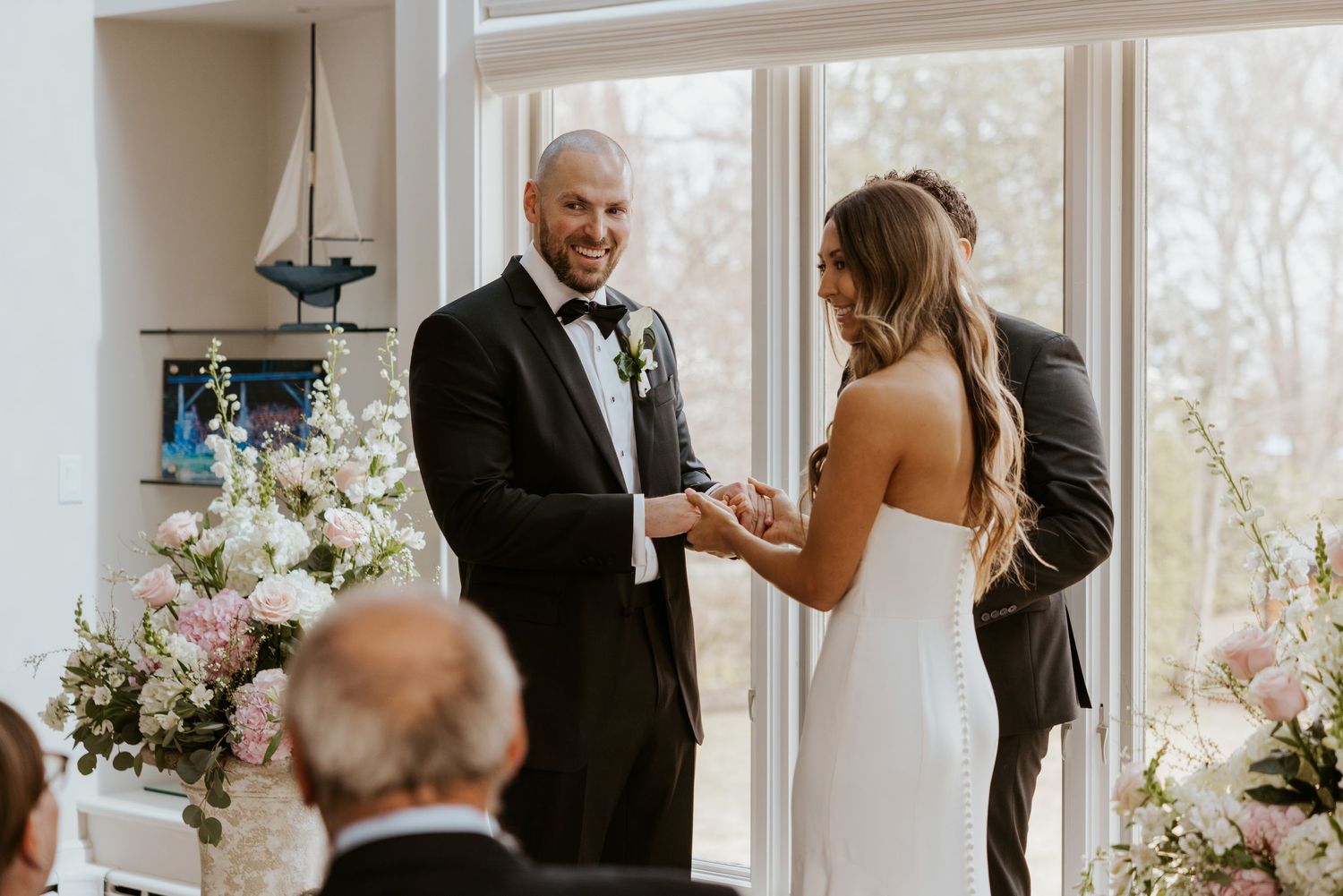 A joyous wedding ceremony takes place in a bright sunlit room decorated with elegant white floral arrangements.