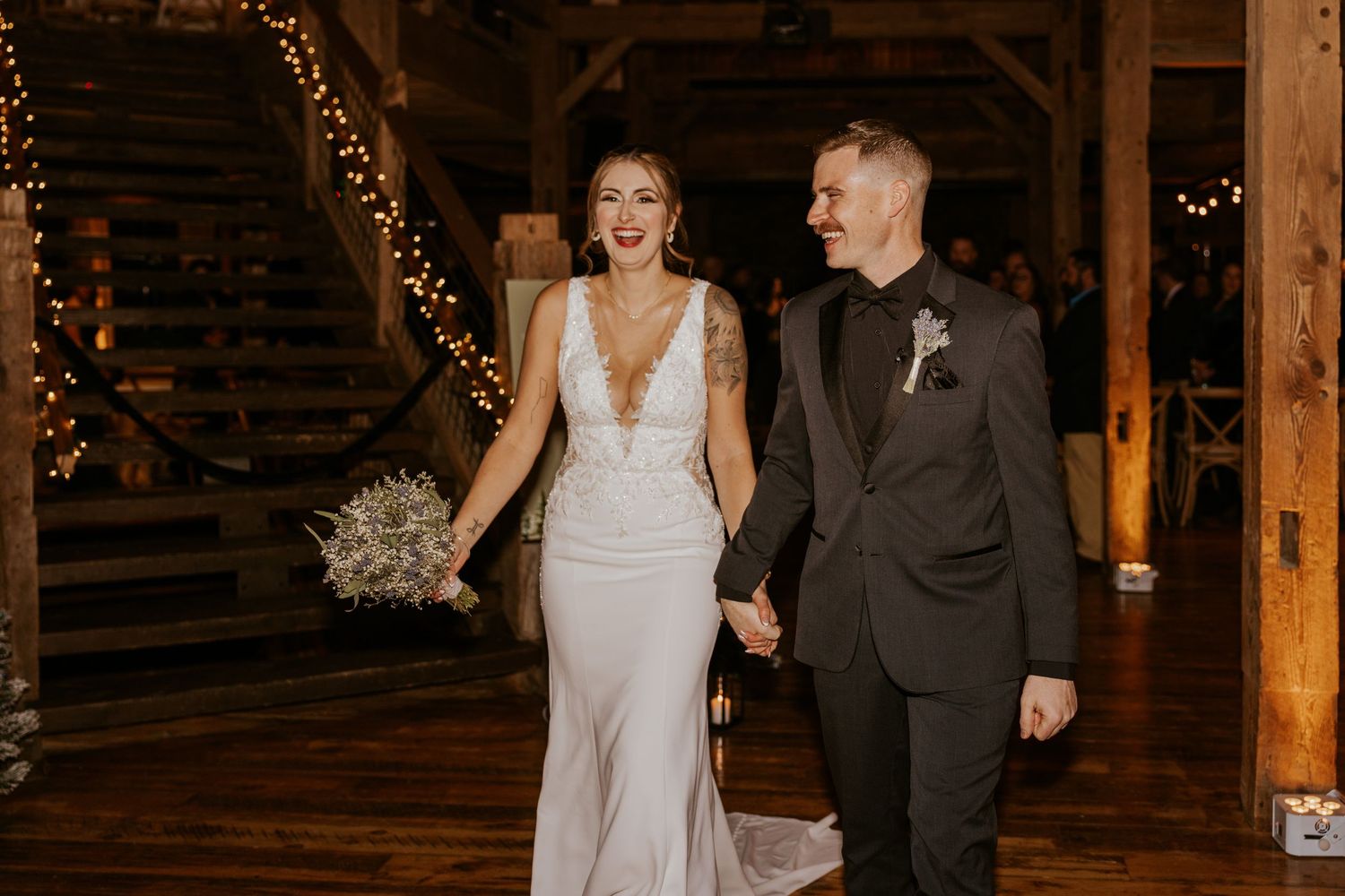 Smiling newlyweds walking hand in hand through warmly lit rustic barn wedding reception venue.
