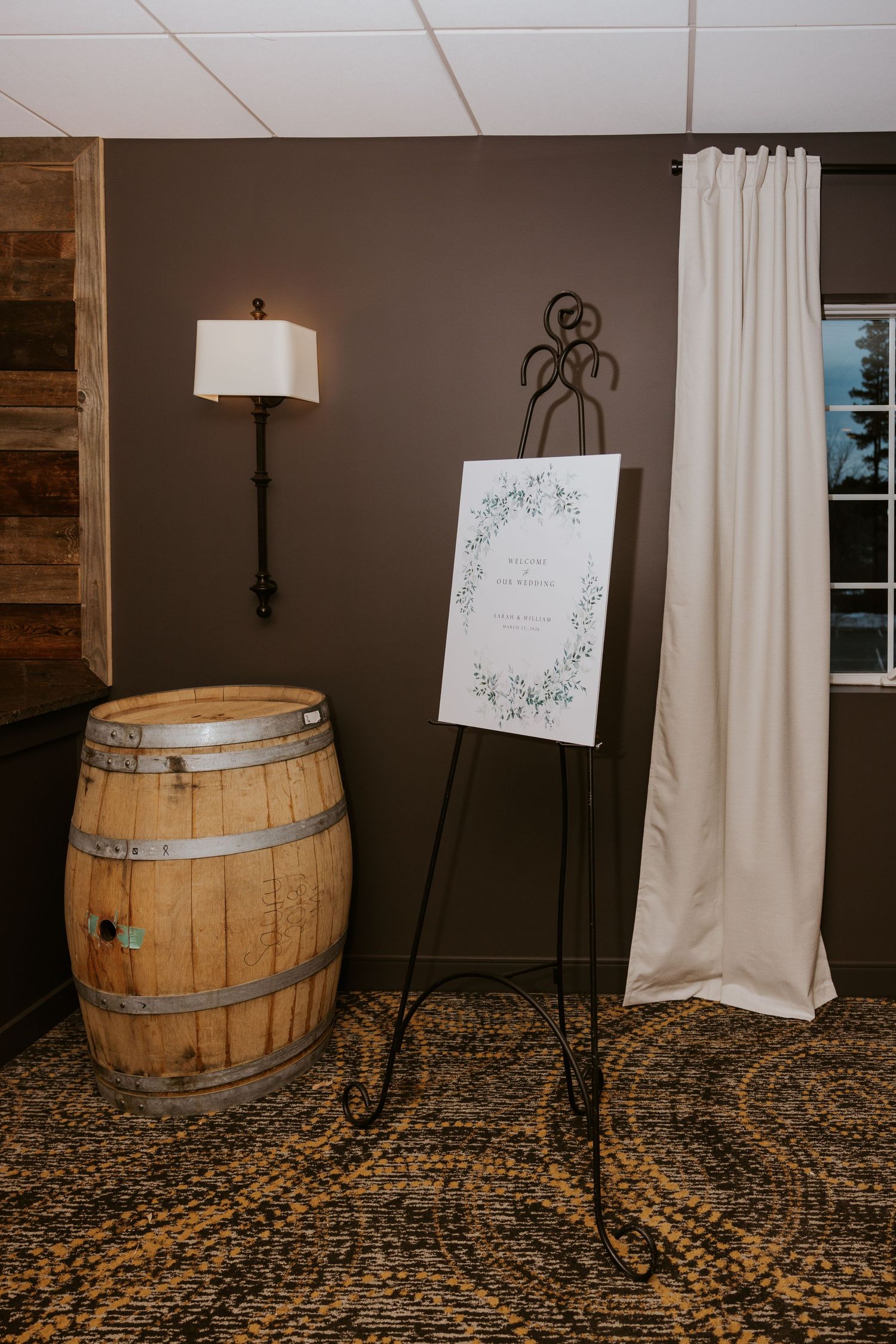 Rustic wine barrels and framed signs decorate an indoor wedding venue with elegant white curtains and carpeted floor.