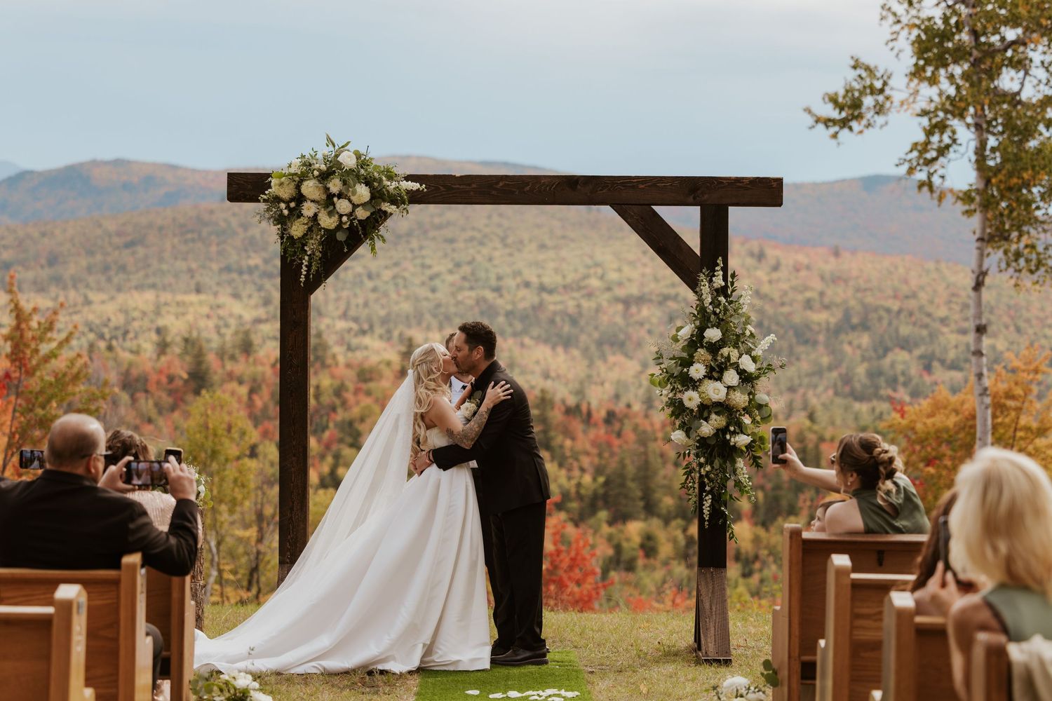 Bride and groom share first kiss under rustic wooden arch with floral arrangements, overlooking colorful autumn mountain foliage.
