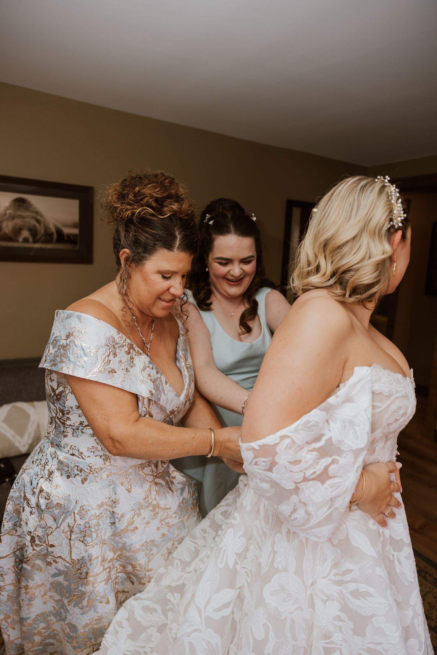 Mother and bridesmaid helping bride button her lace wedding gown before the ceremony.