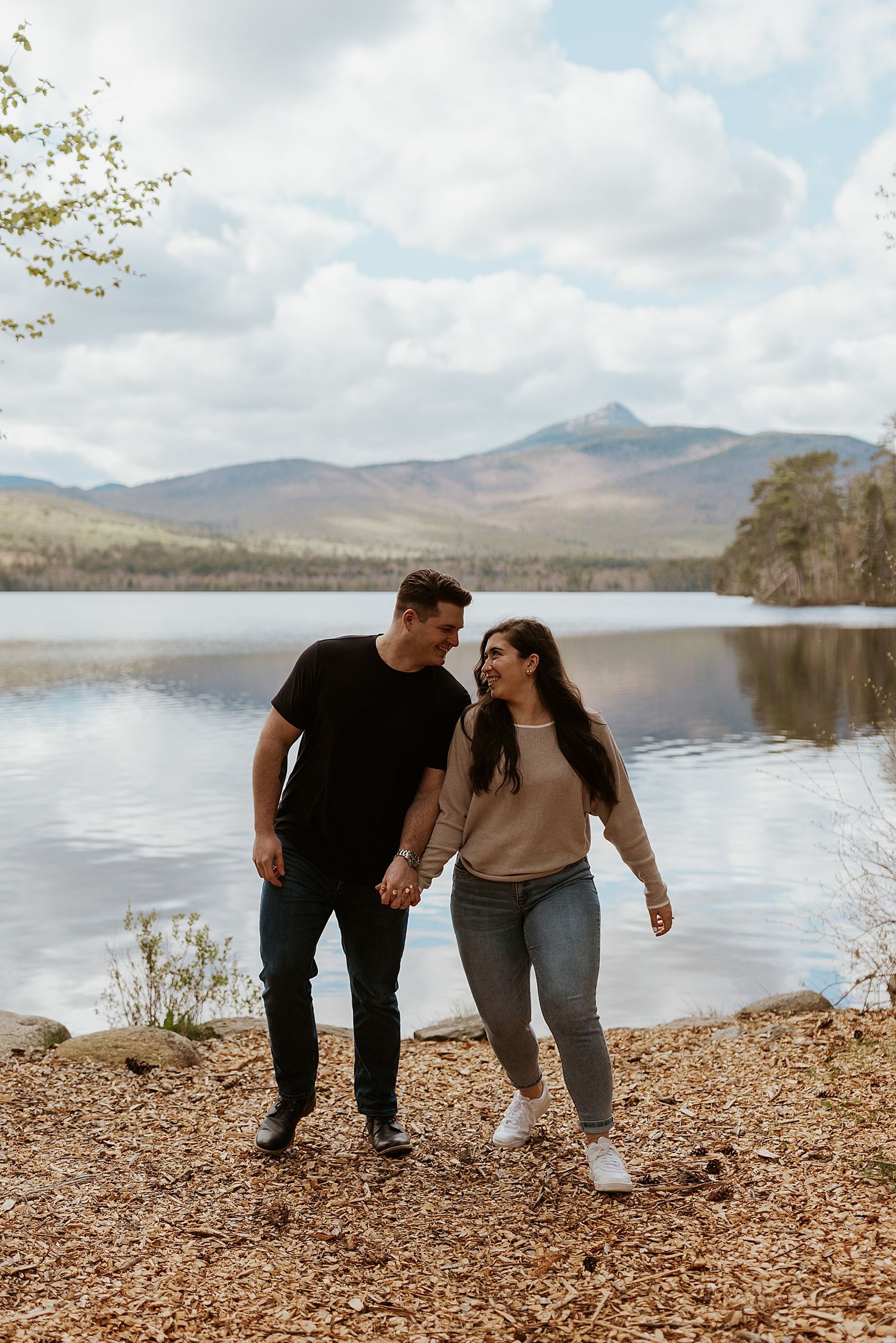 Couple holding hands and smiling at each other by a scenic lake with mountains in the background.