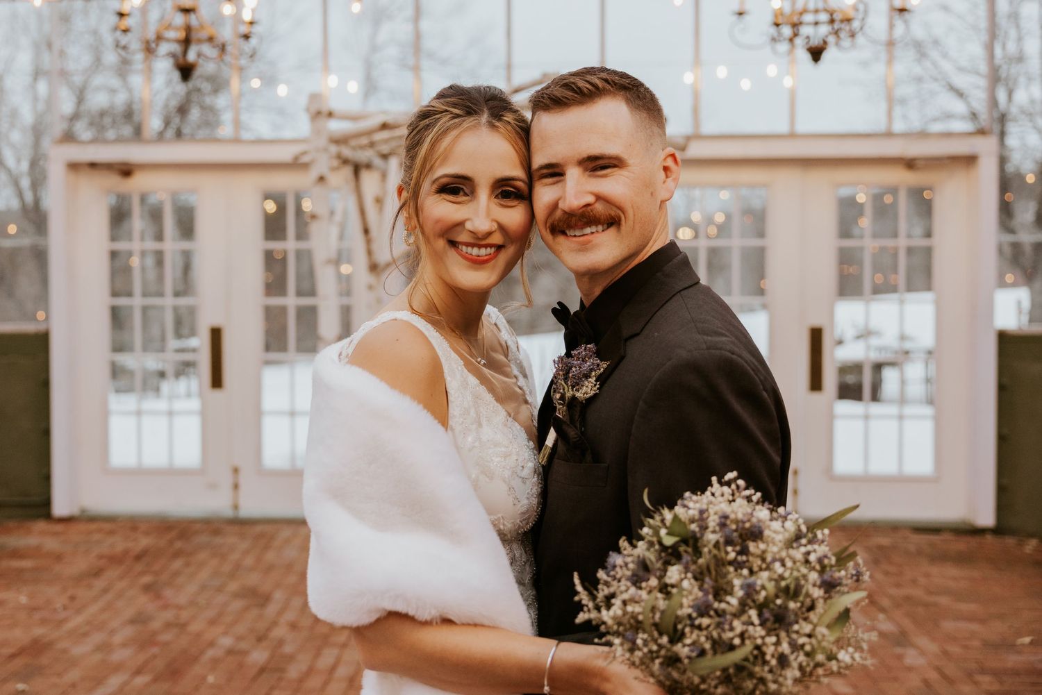 Bride and groom smiling together outside elegant greenhouse wedding venue in winter, bride holding bouquet.