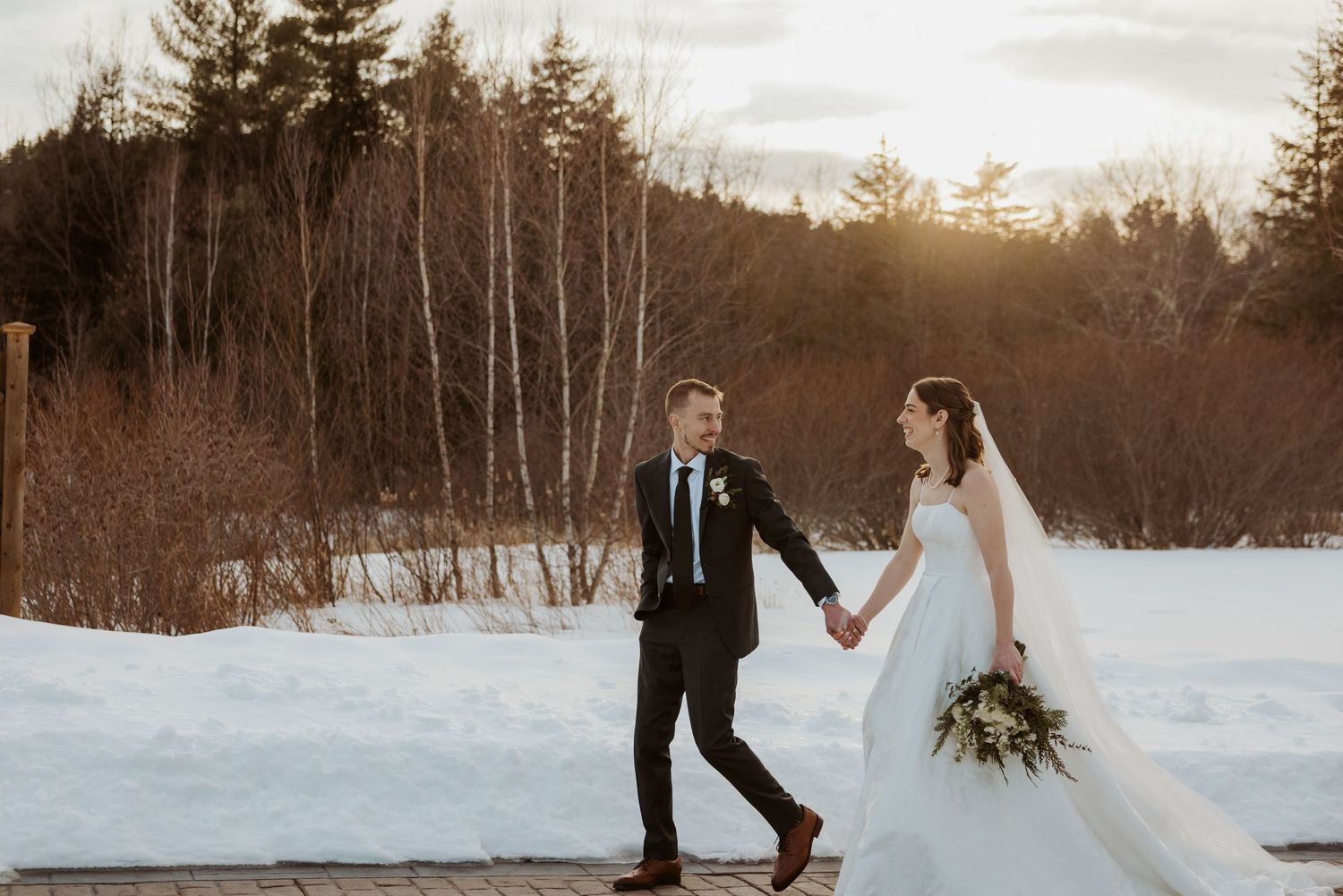 Couple in wedding attire walks together in a snowy winter landscape at sunset with evergreen trees in background.
