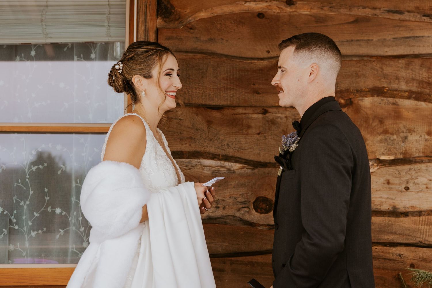 Bride in white gown with fur wrap reading vows to groom inside a cozy log cabin wedding venue.