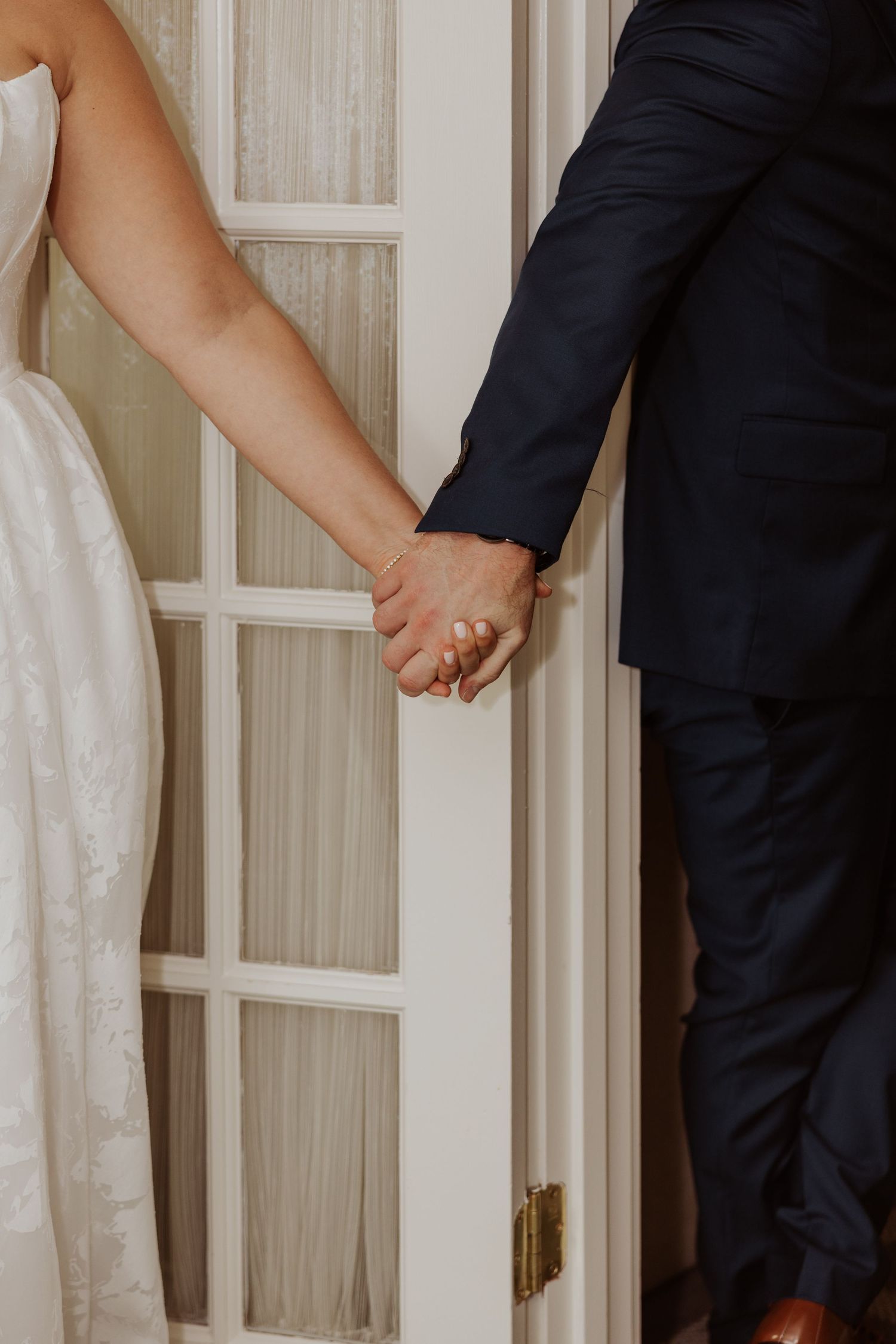 A bride in lace and groom in navy suit hold hands through a glass-paned door before the ceremony.