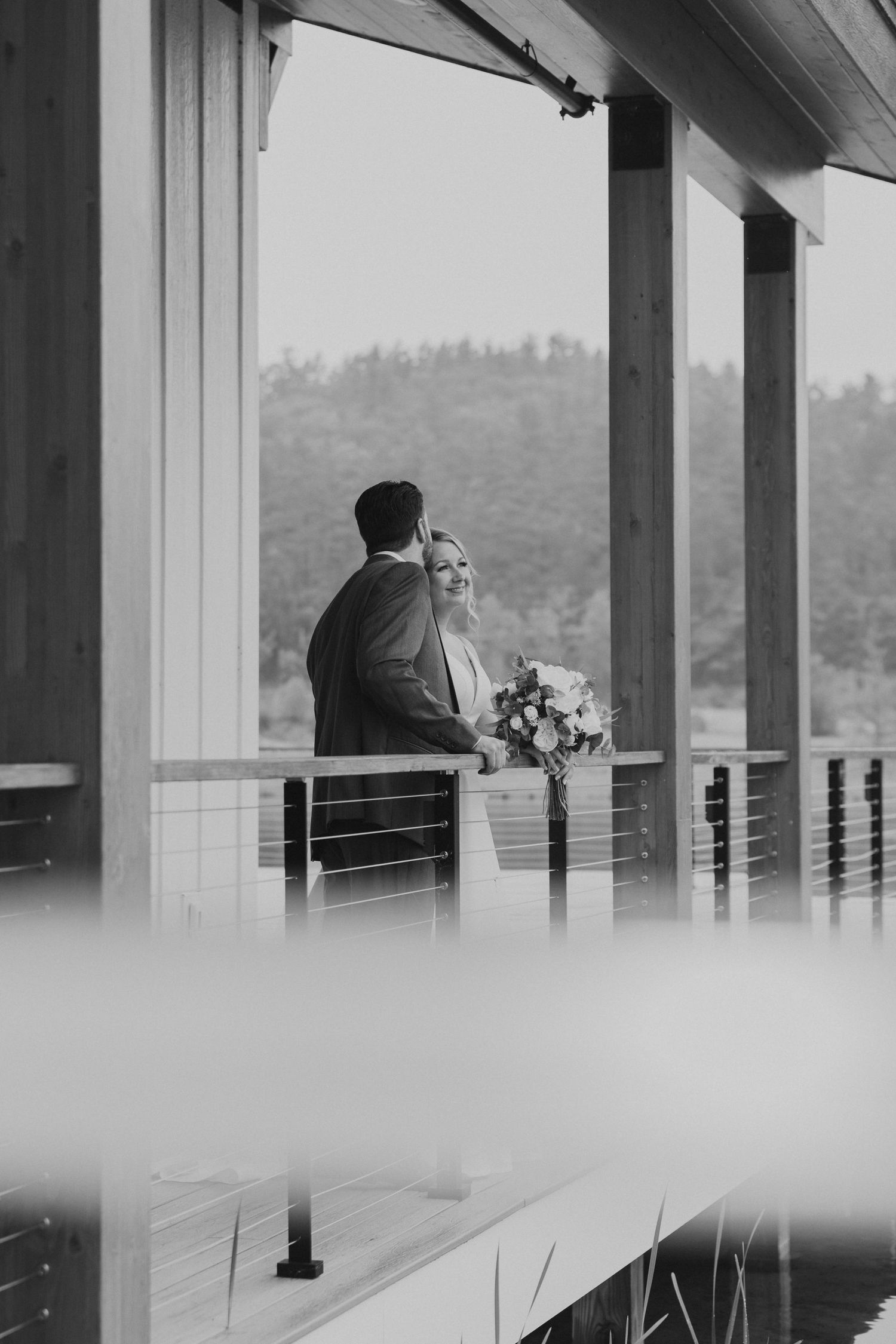 Black and white photo of bride and groom standing on a balcony overlooking a forested landscape.