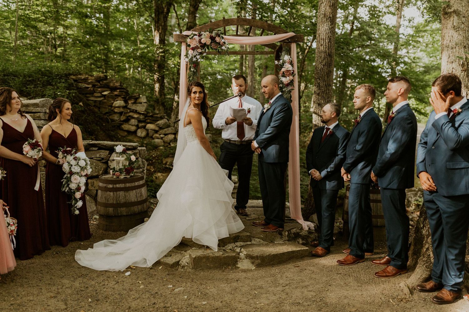 Forest wedding ceremony with guests seated on wooden benches under a canopy of trees.