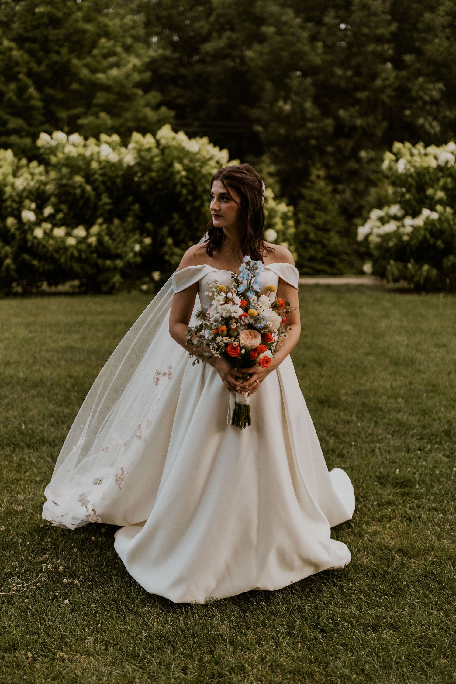 Bride in off-shoulder wedding gown holds colorful bouquet in garden setting with white hydrangeas.