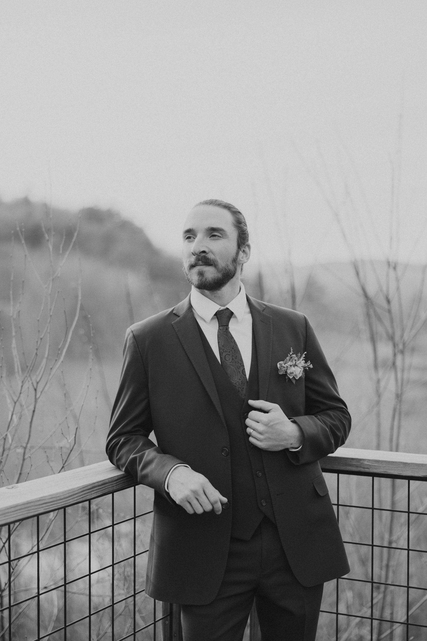 Dapper groom in dark suit with boutonniere poses on outdoor railing in black and white photo.