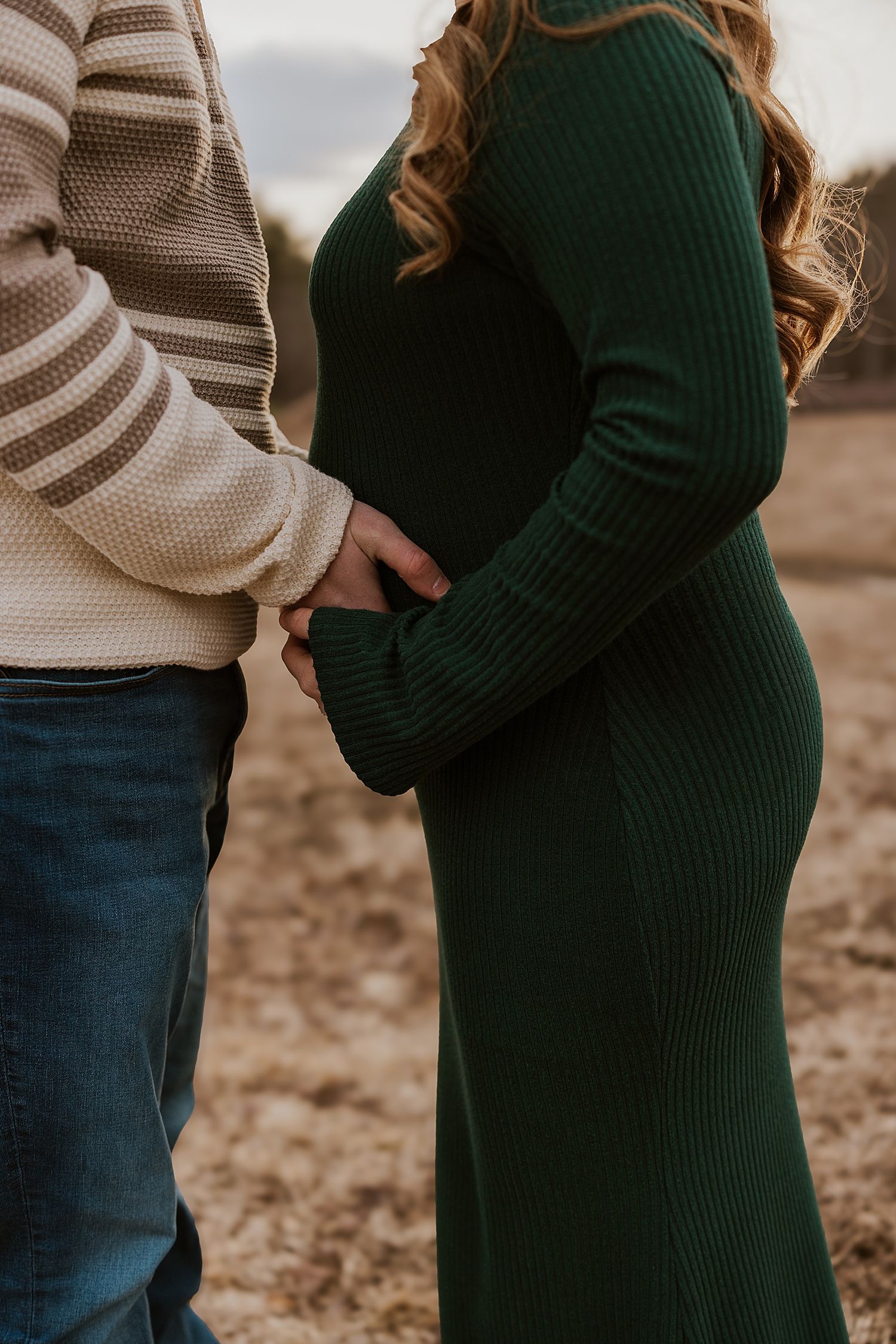 Couple holding pregnant belly outdoors in a field, woman wearing green ribbed maternity dress, man in striped sweater.