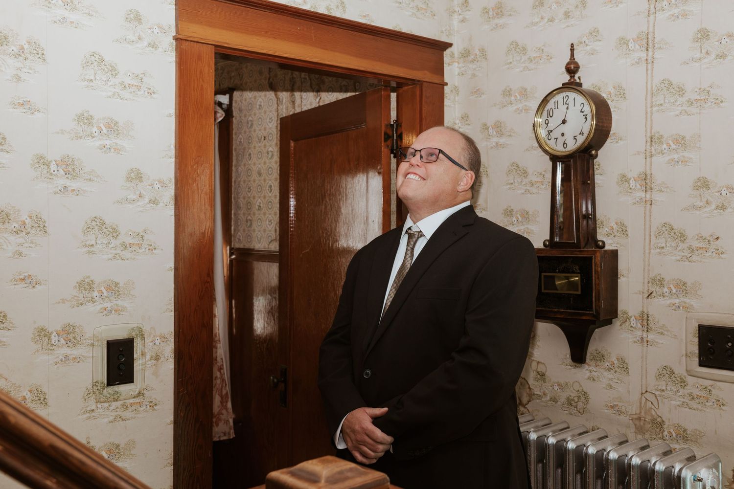 Man in black suit looking up in vintage room with wallpaper, wooden door, and antique pendulum clock.