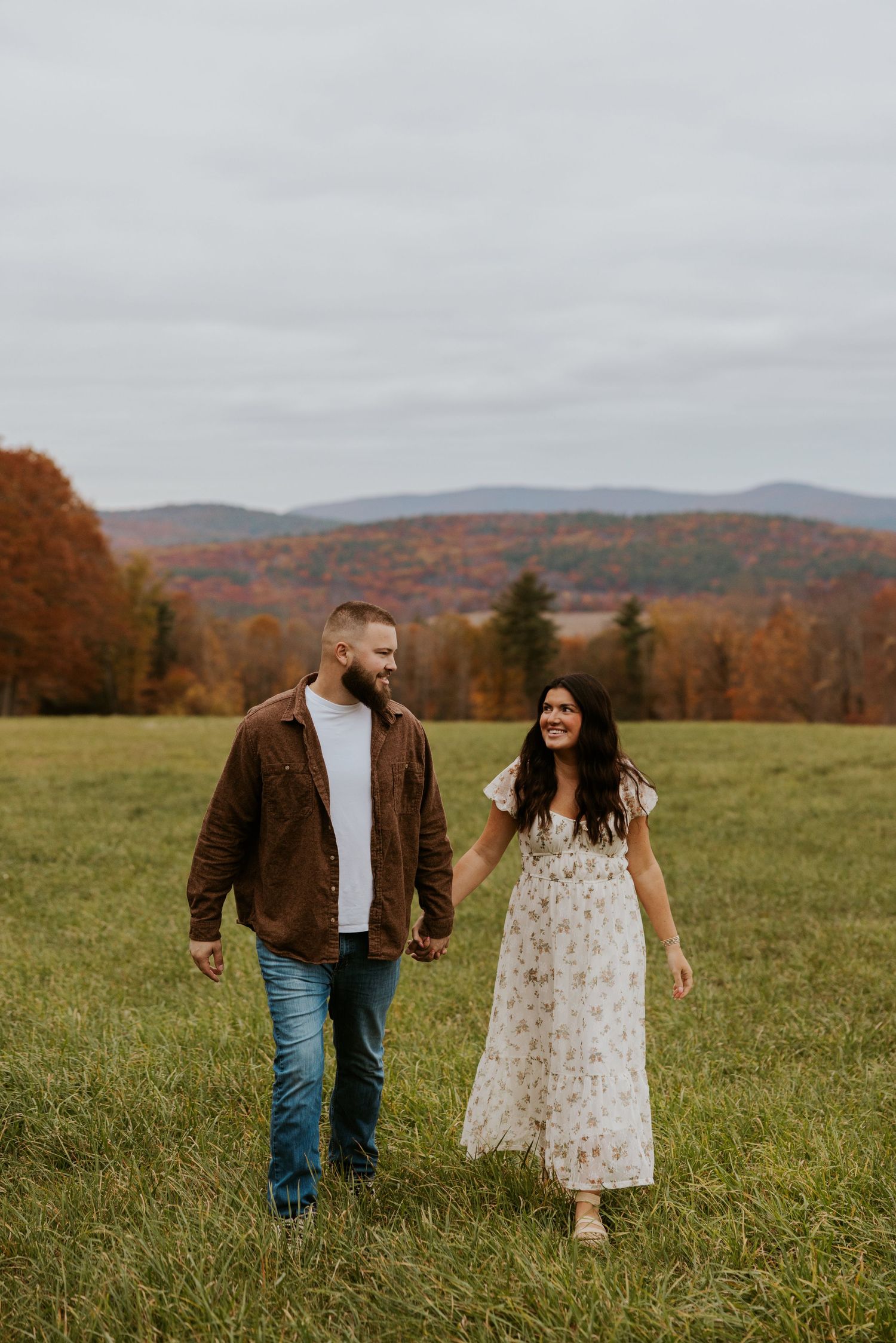 Smiling couple walks through green autumn field with colorful fall mountain foliage.