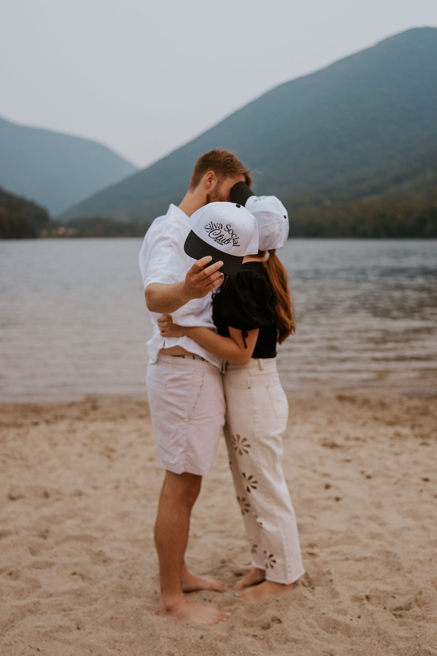 Couple kissing on a sandy beach lakeside with lush green mountains in the background.