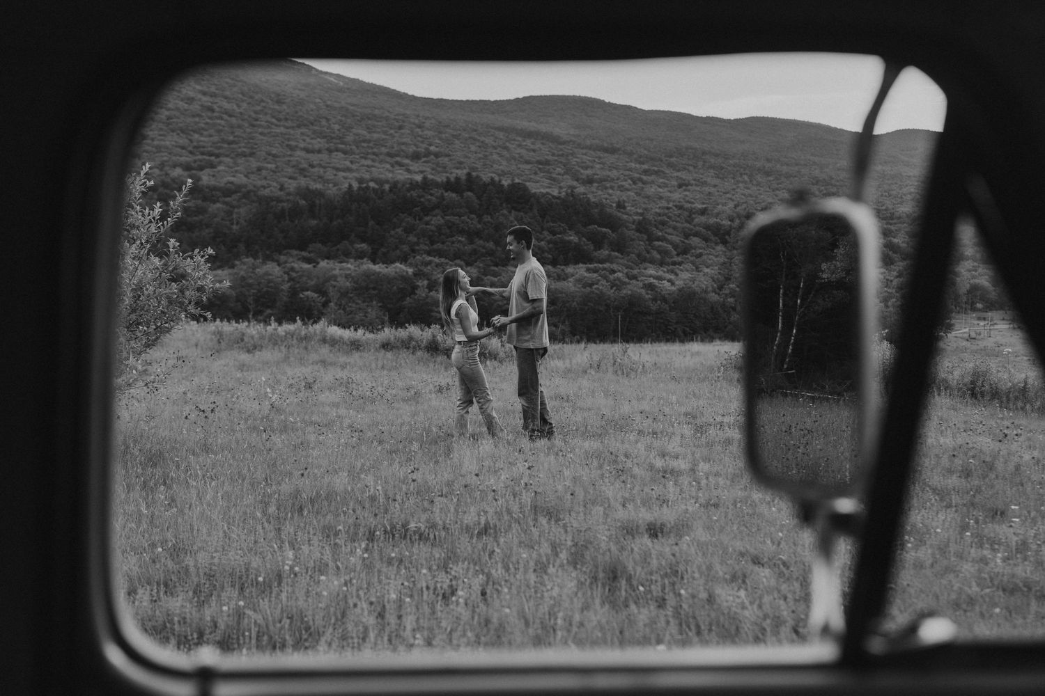 Black and white photo of a couple holding hands in a meadow, framed by a vintage truck window.