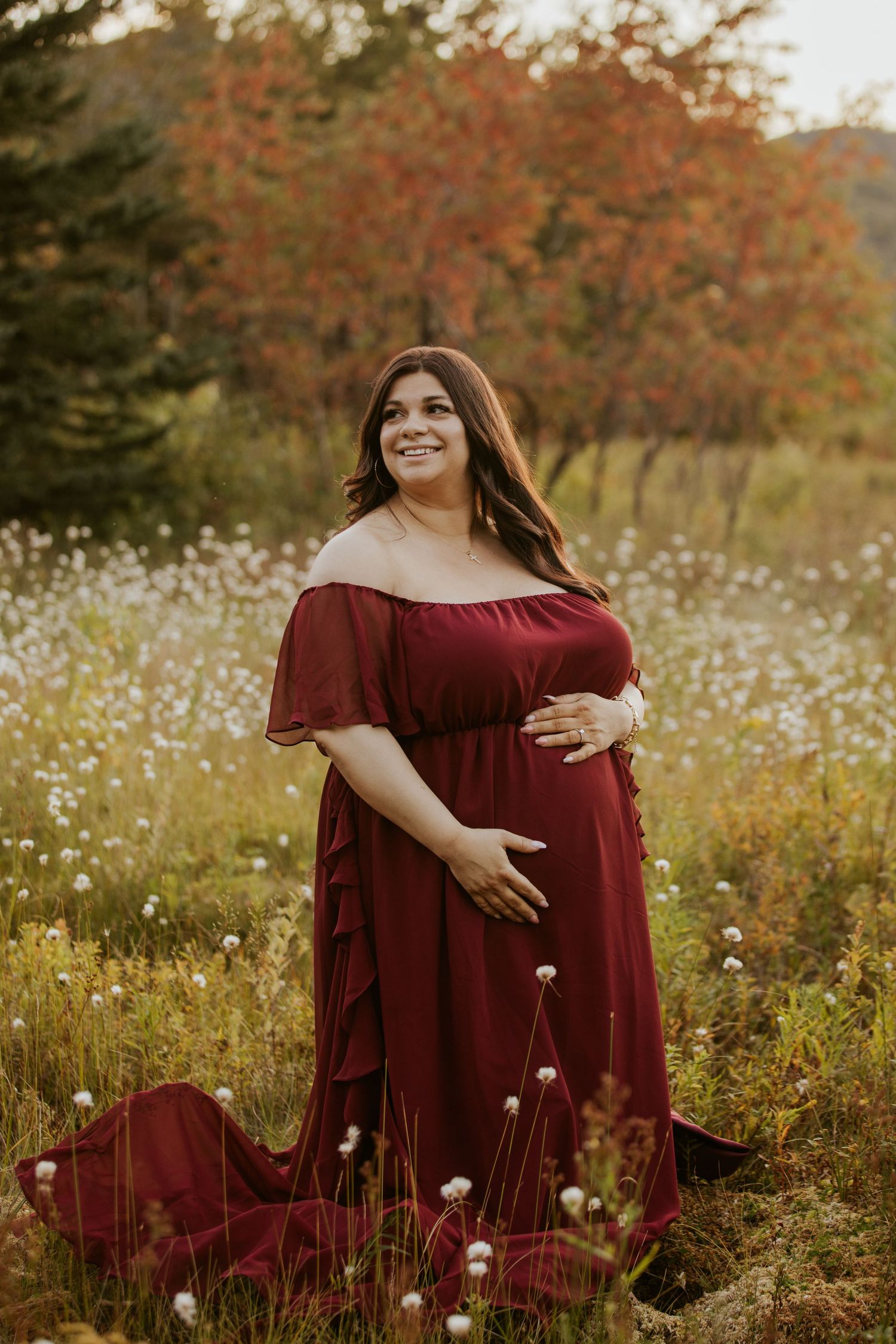 Pregnant woman in burgundy off-shoulder gown standing in wildflower field with autumn foliage background.