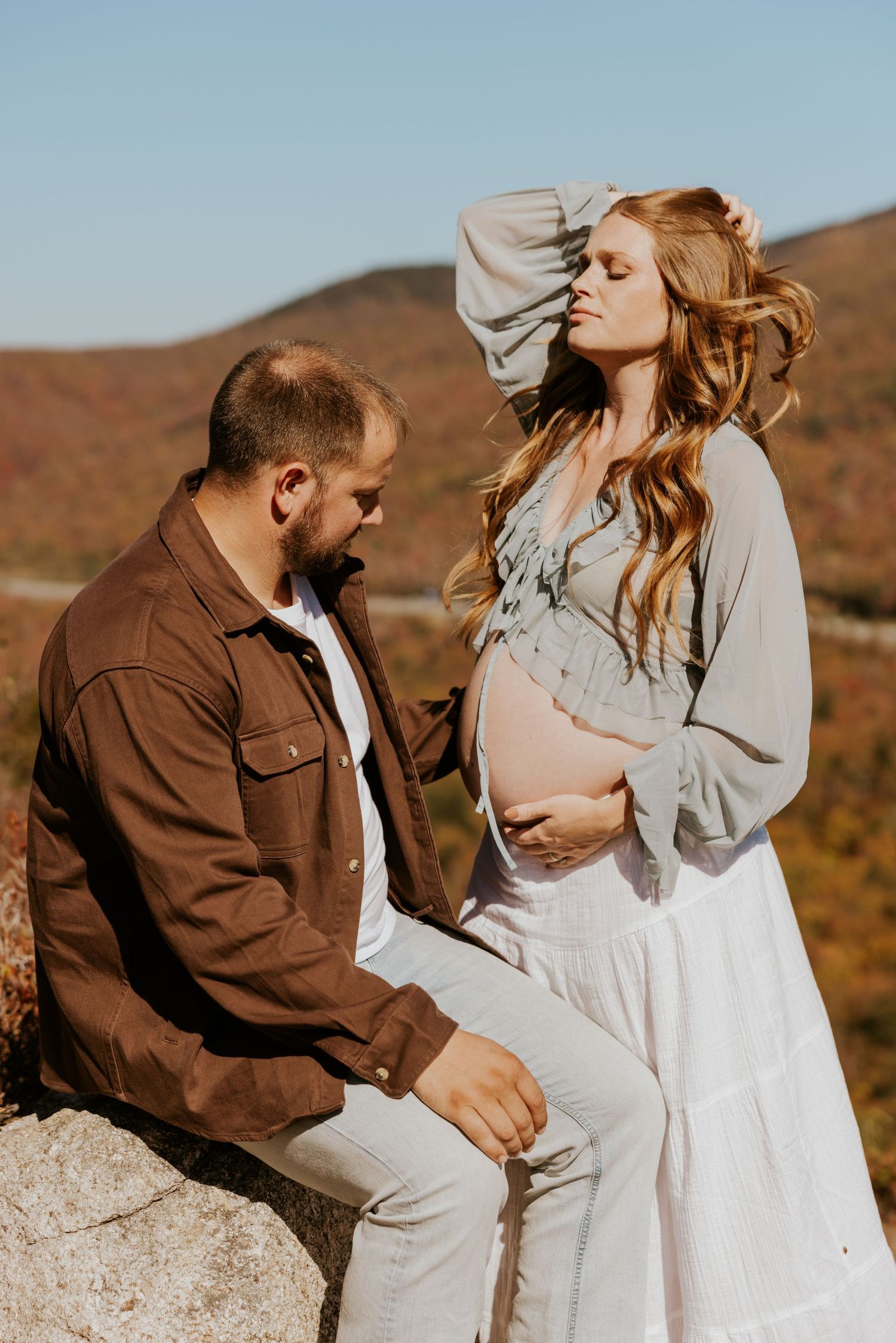 Expectant couple on rocky summit, woman in white skirt cradling bump with fall mountains backdrop.