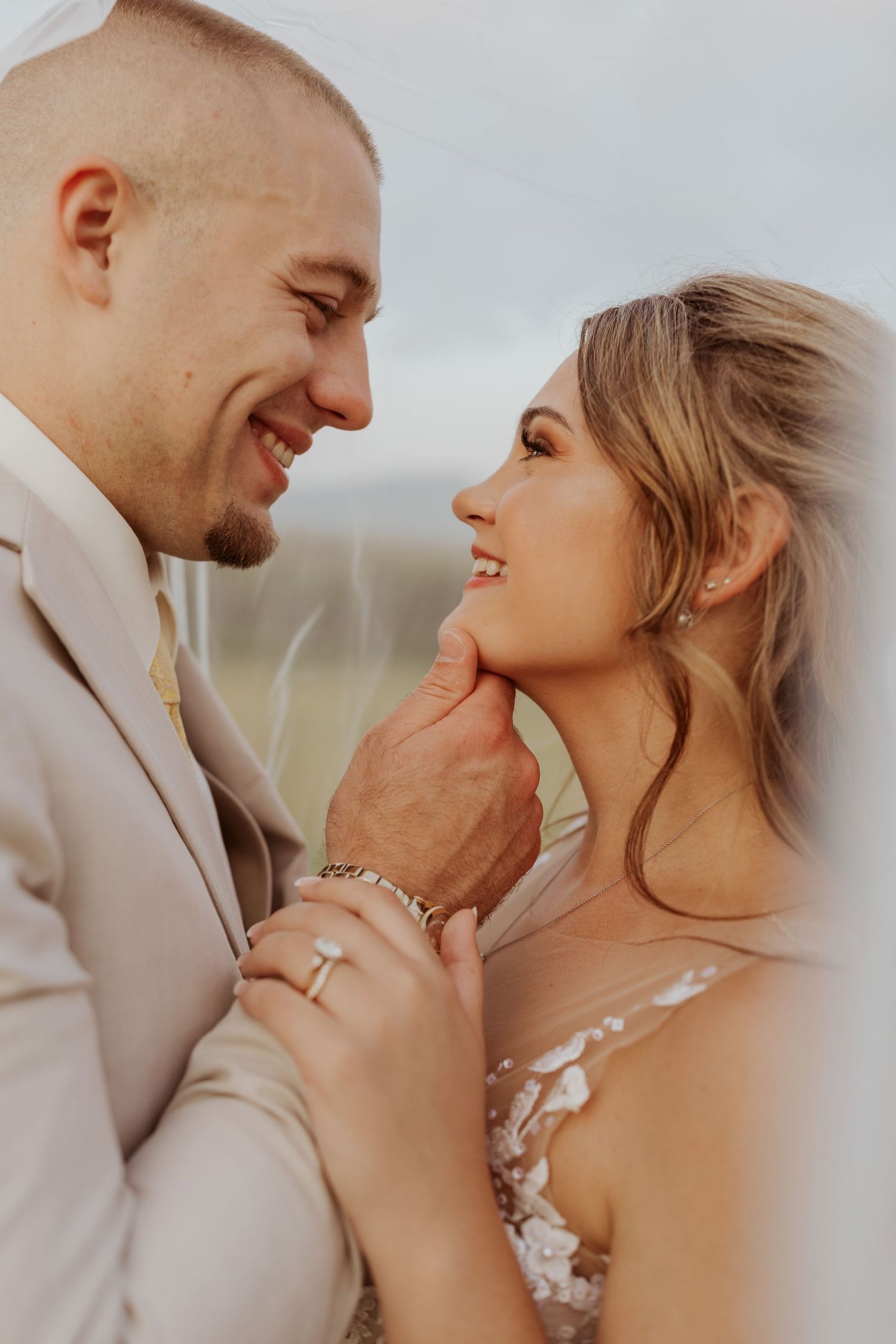 Smiling groom gently holds brides chin as they gaze at each other on their wedding day outdoors.