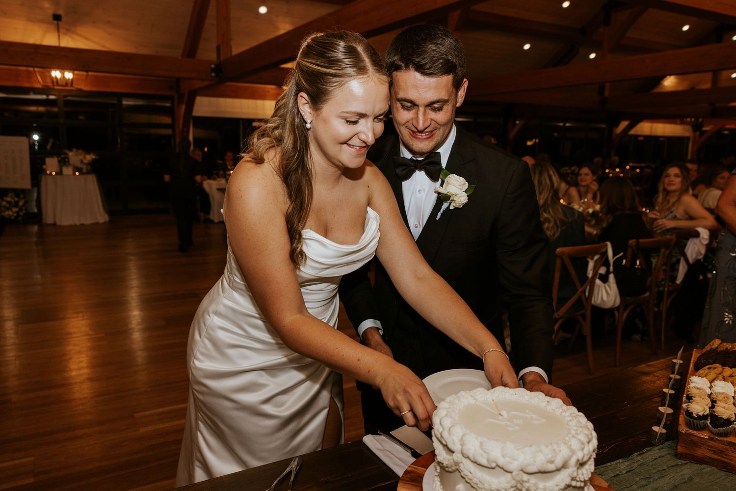 Bride and groom smiling while cutting their white wedding cake together at their indoor reception venue.