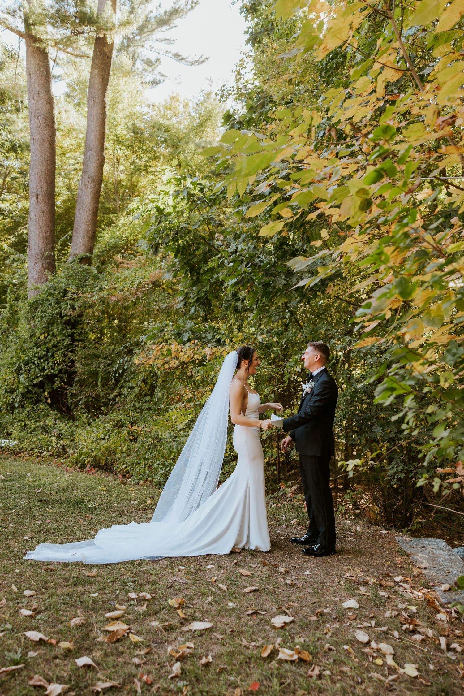 Bride and groom hold hands in full-length wedding attire surrounded by colorful autumn foliage.