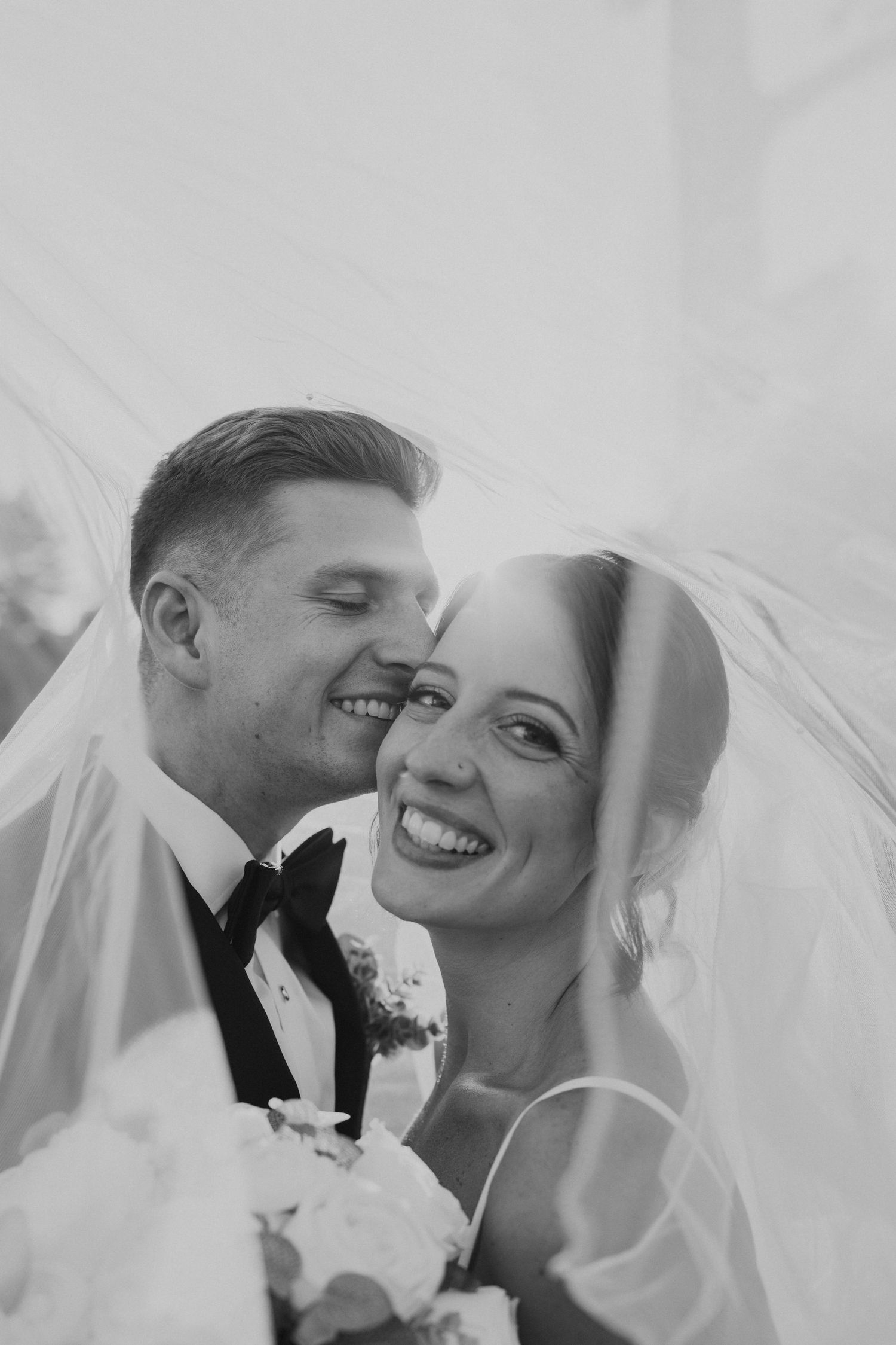 Black and white portrait of smiling bride and groom nestled under wedding veil.