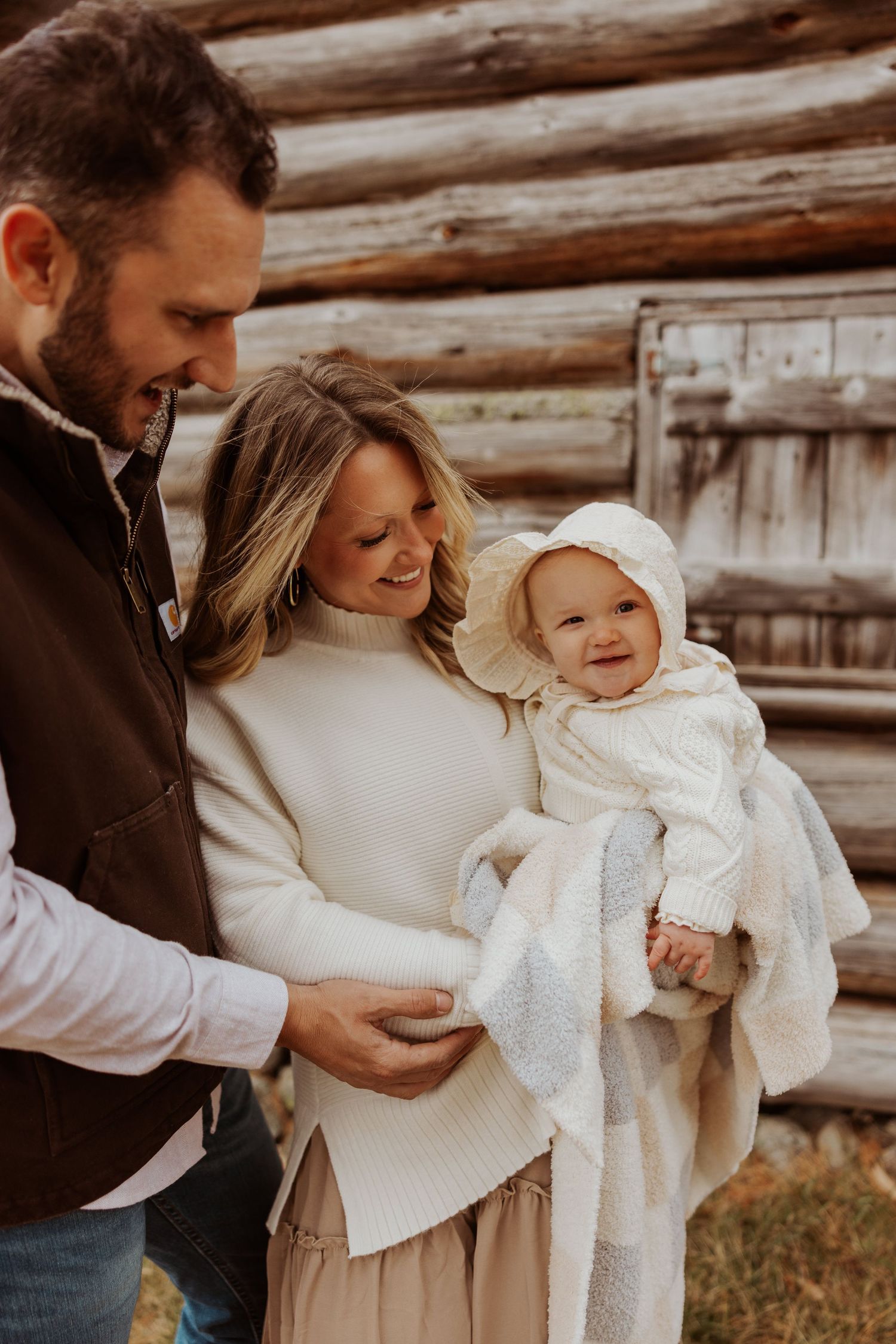 Smiling family with father, mother in white sweater, and baby in bonnet near rustic log wall.