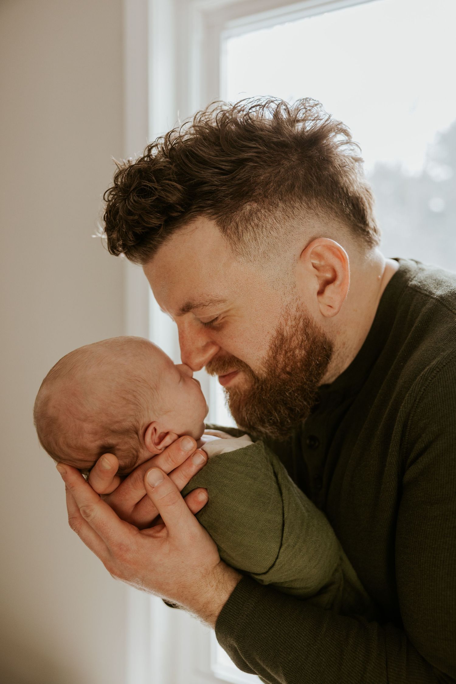 Bearded father tenderly touching foreheads with sleeping newborn baby held in hands by window.