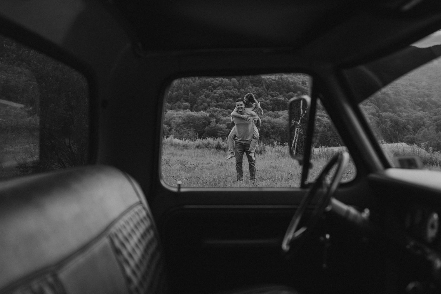 Black and white view through a vintage truck window of a man giving a woman a piggyback ride.