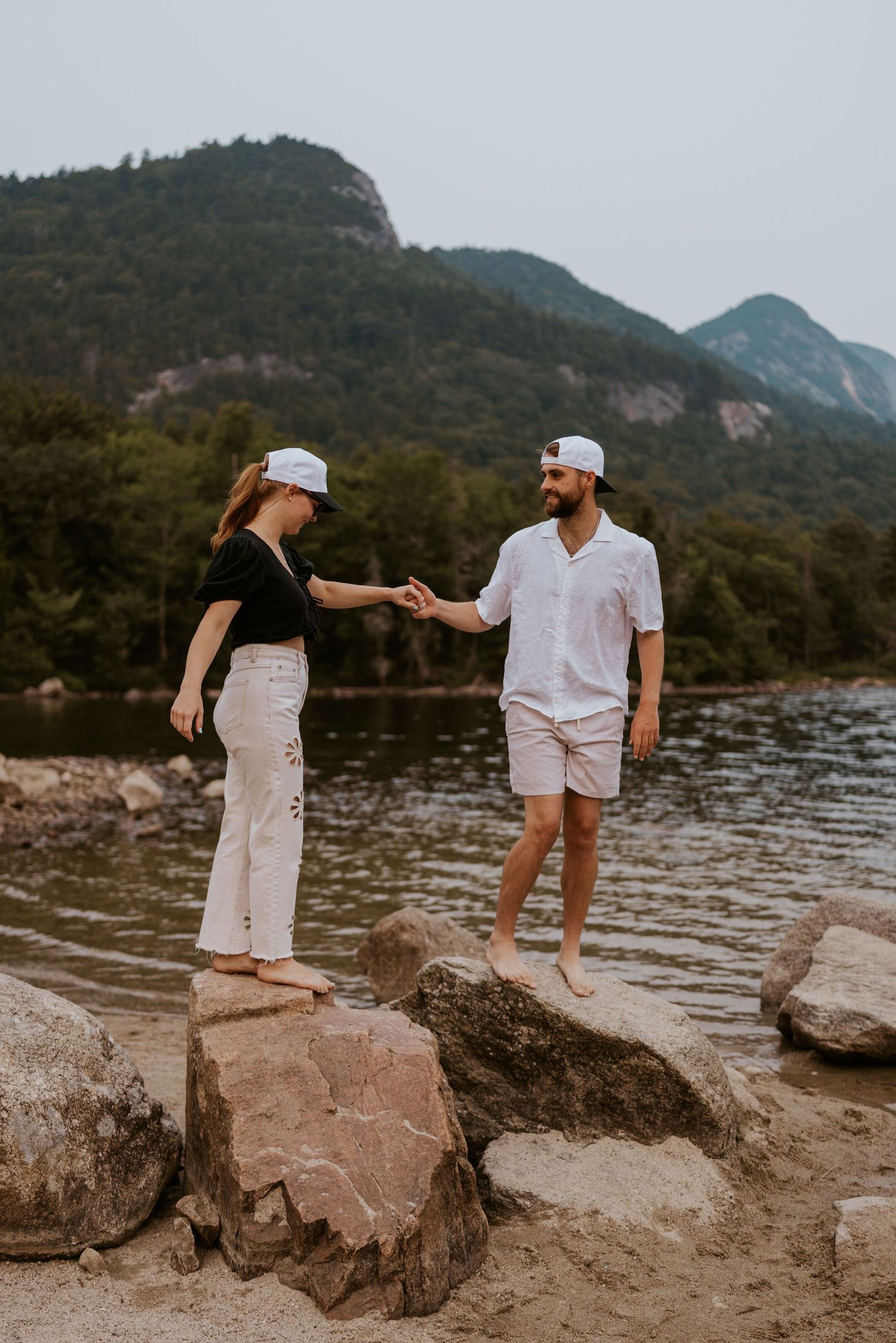 Couple holding hands while standing on rocks by a mountain lake at dusk.
