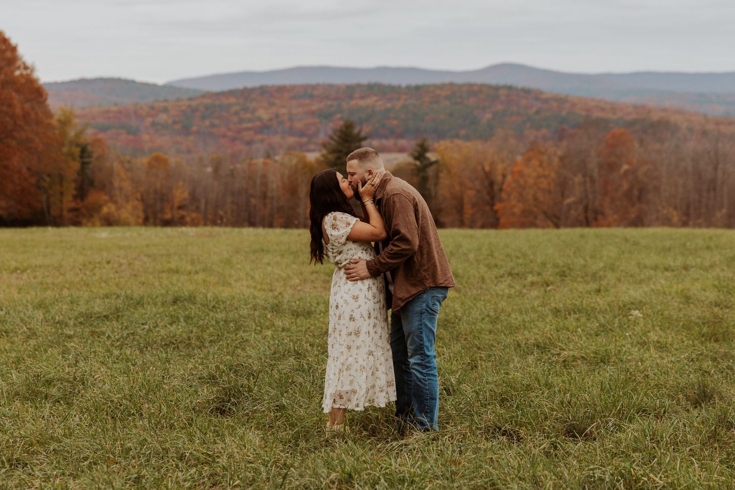 Couple sharing a romantic kiss in an open green field with colorful autumn mountain foliage in the background.