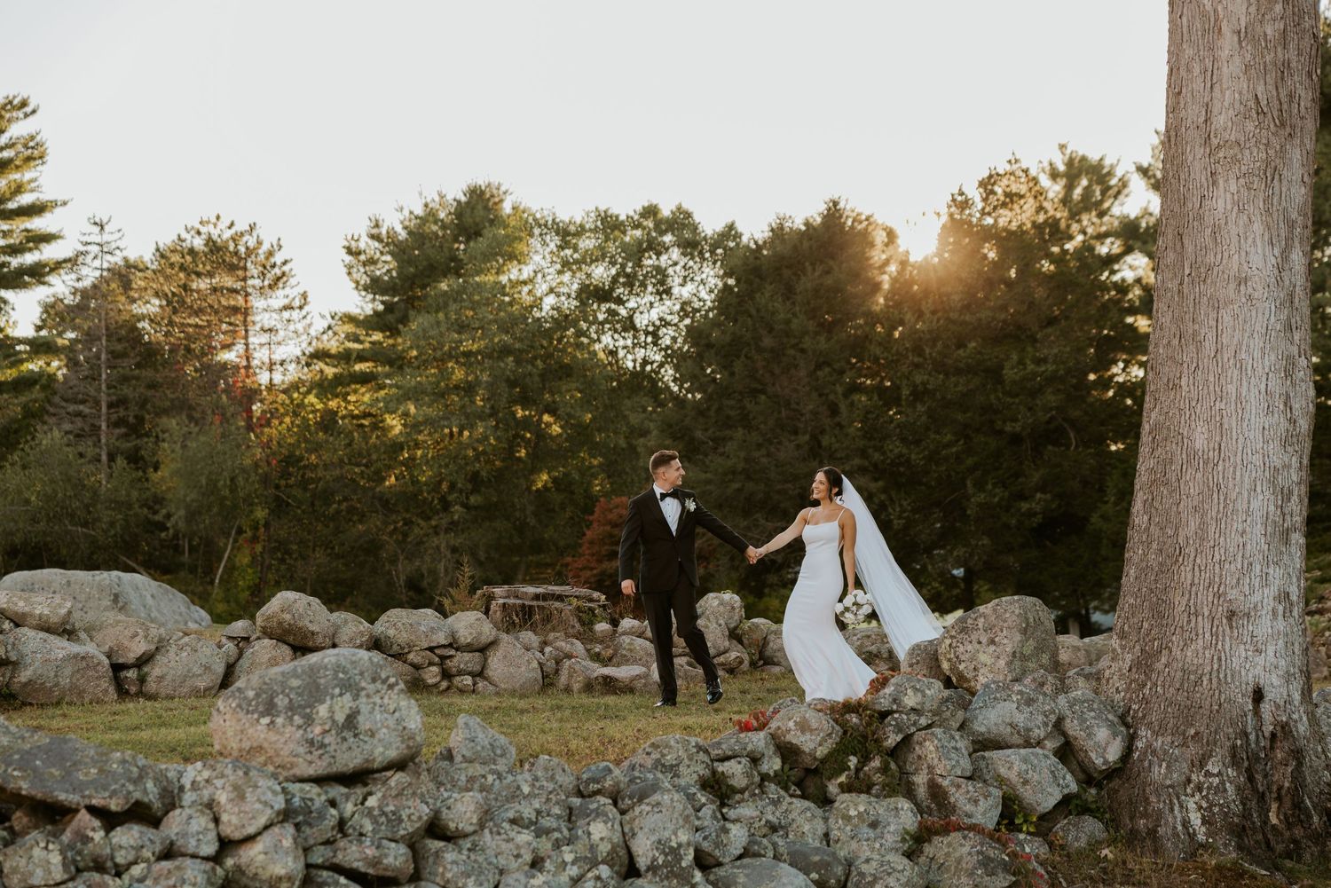 Bride and groom hold hands walking among rustic stone walls at sunset.