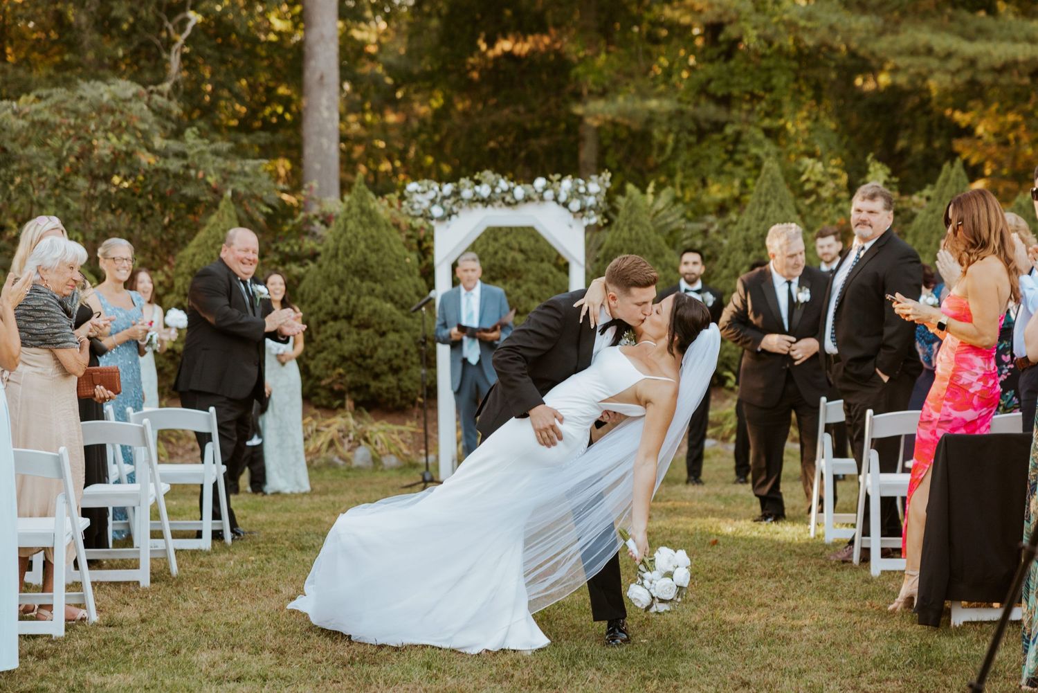 Groom dips bride for kiss while wedding guests cheer along decorated outdoor ceremony aisle.