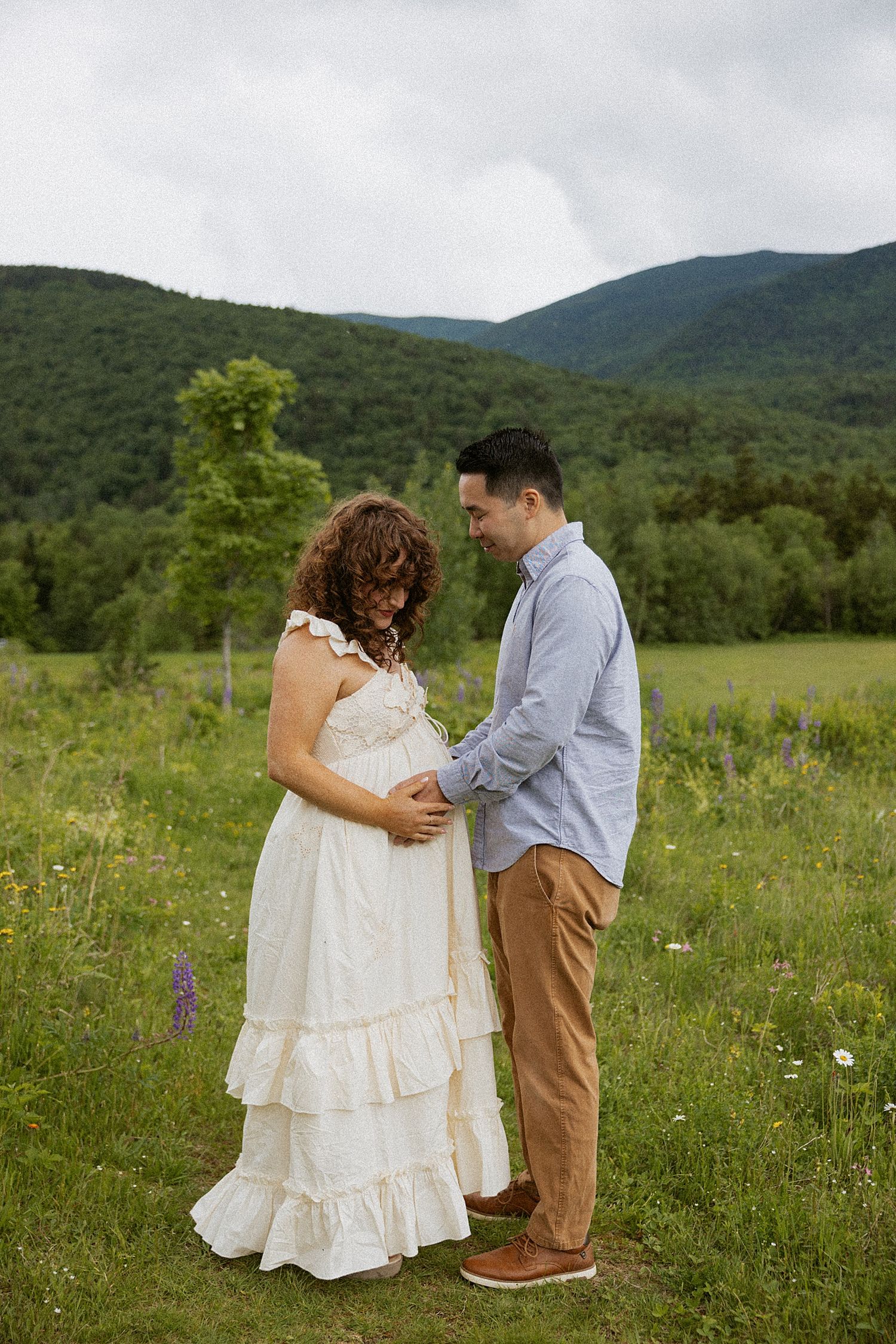 Pregnant couple holding hands in a green meadow with mountain backdrop in New Hampshire, taking motherhood photos