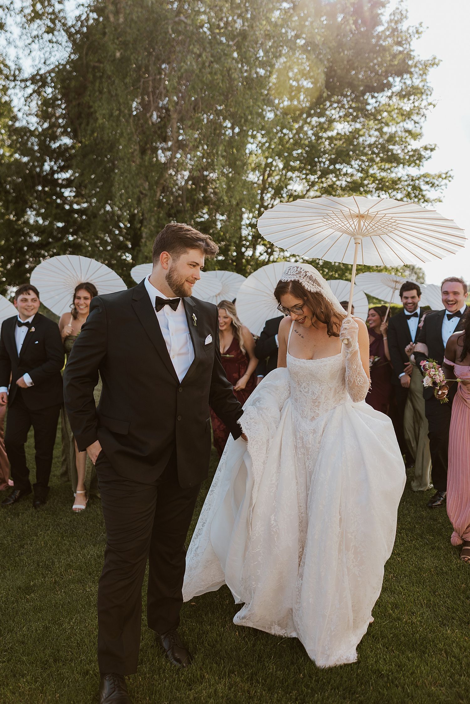 Bride and groom walk together outdoors as wedding guests hold white parasols in golden sunlight at a New Hampshire wedding