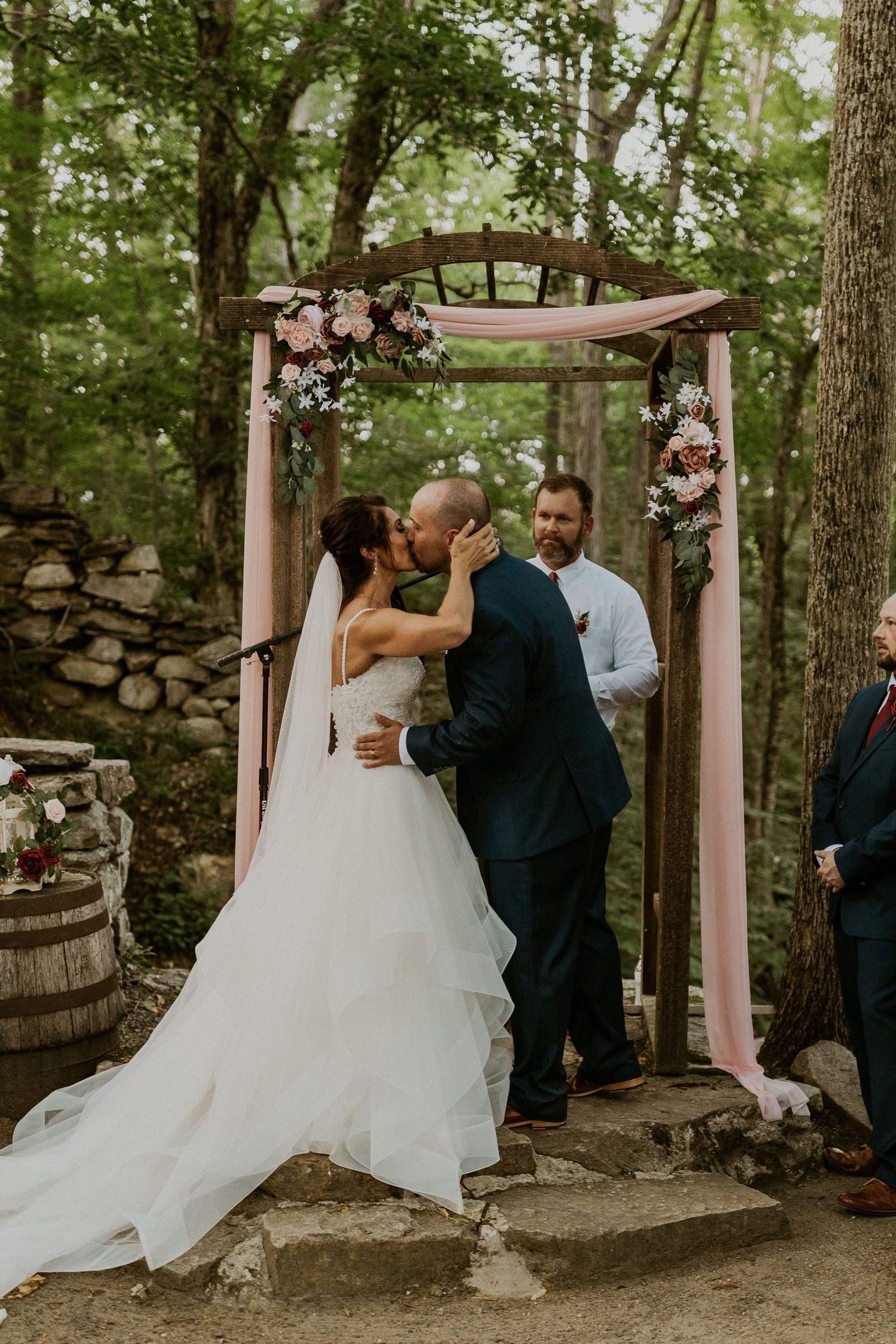 Wedding couple shares an intimate moment under trees with guests looking on.
