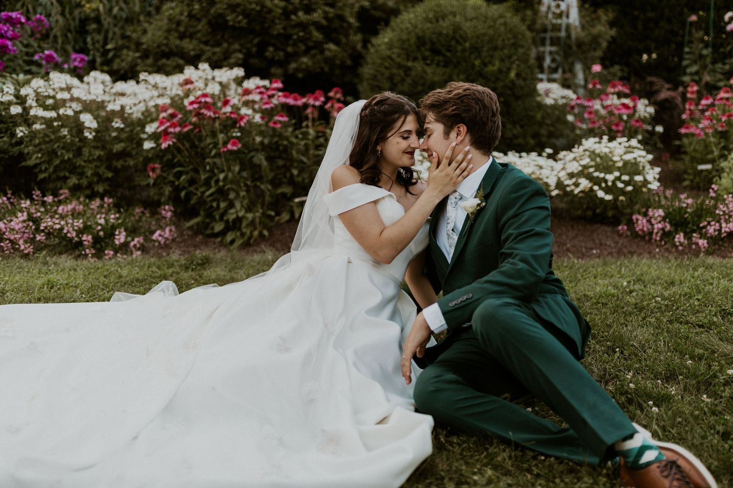 Wedding couple shares intimate moment on lawn surrounded by pink and white flowering bushes.
