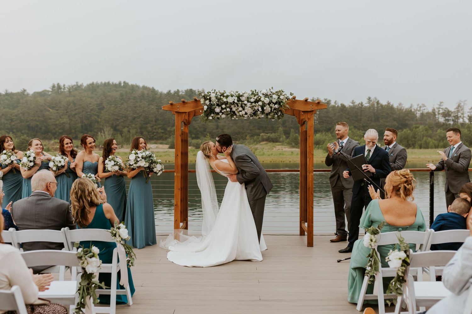 Bride and groom share first kiss at outdoor lakeside ceremony beneath floral-adorned wooden arch.