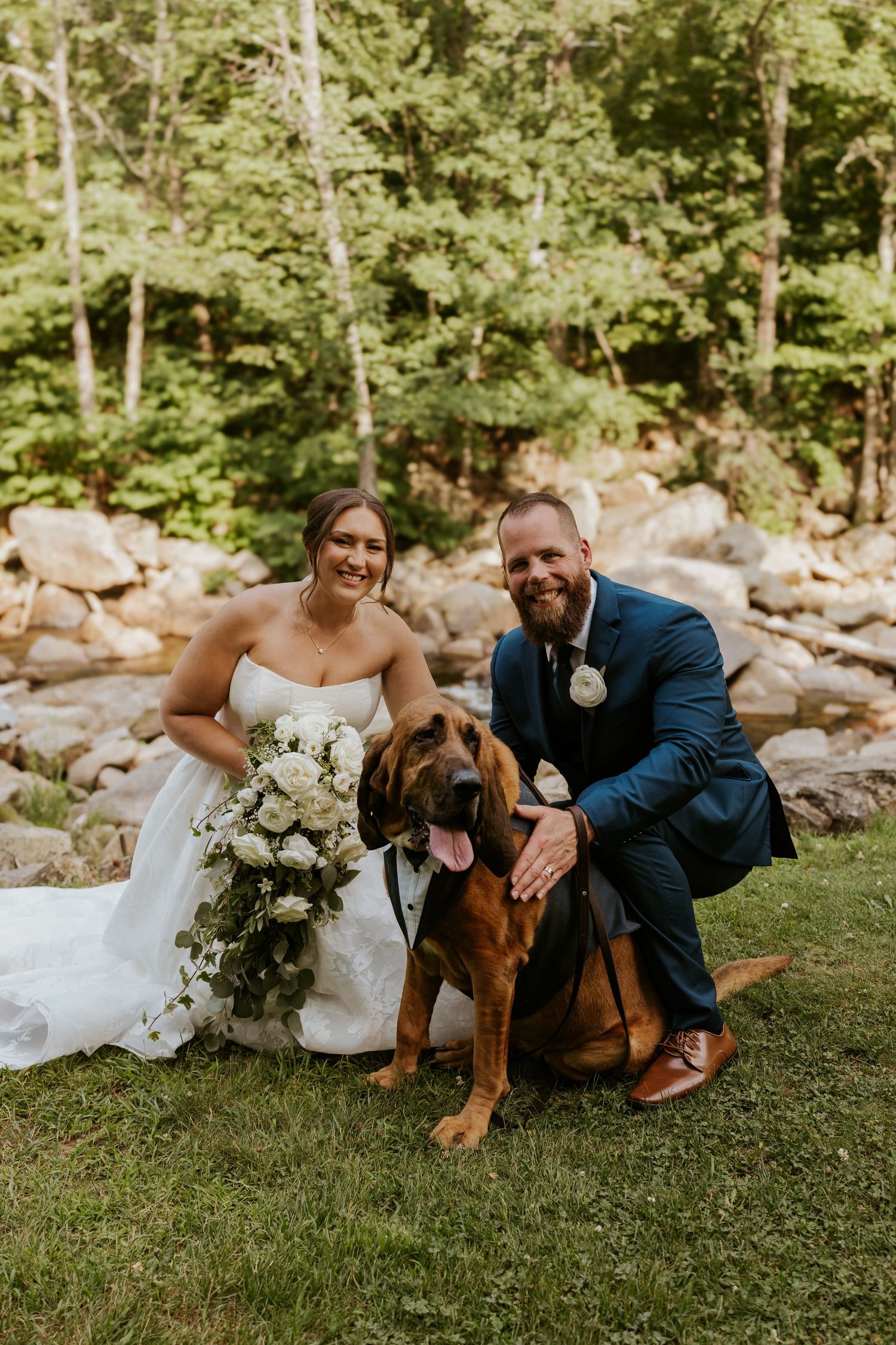 A smiling bride and groom pose with their large brown dog in front of a rocky wooded backdrop.