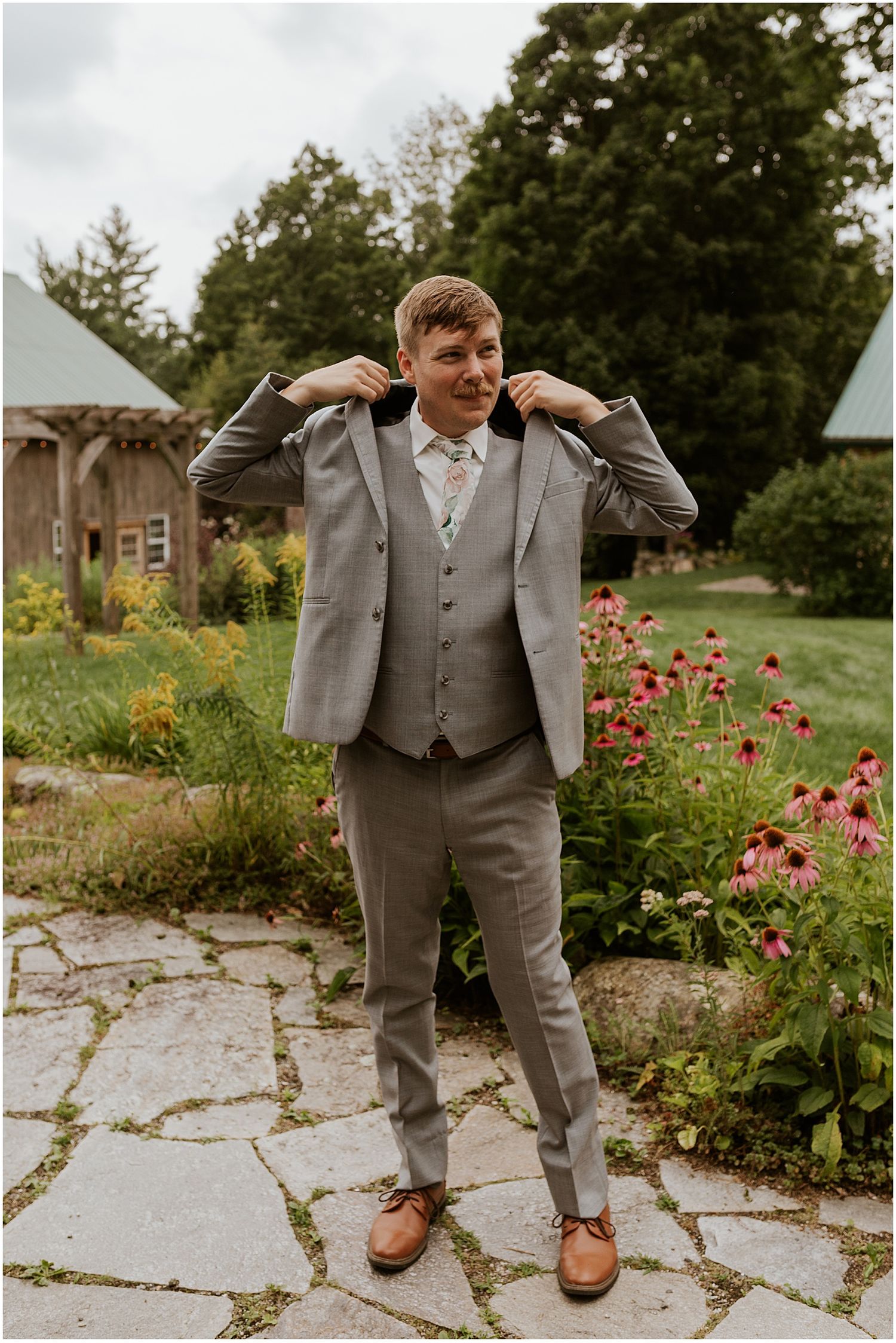A groom in a light gray suit adjusts his collar while standing on a stone patio surrounded by flowers.