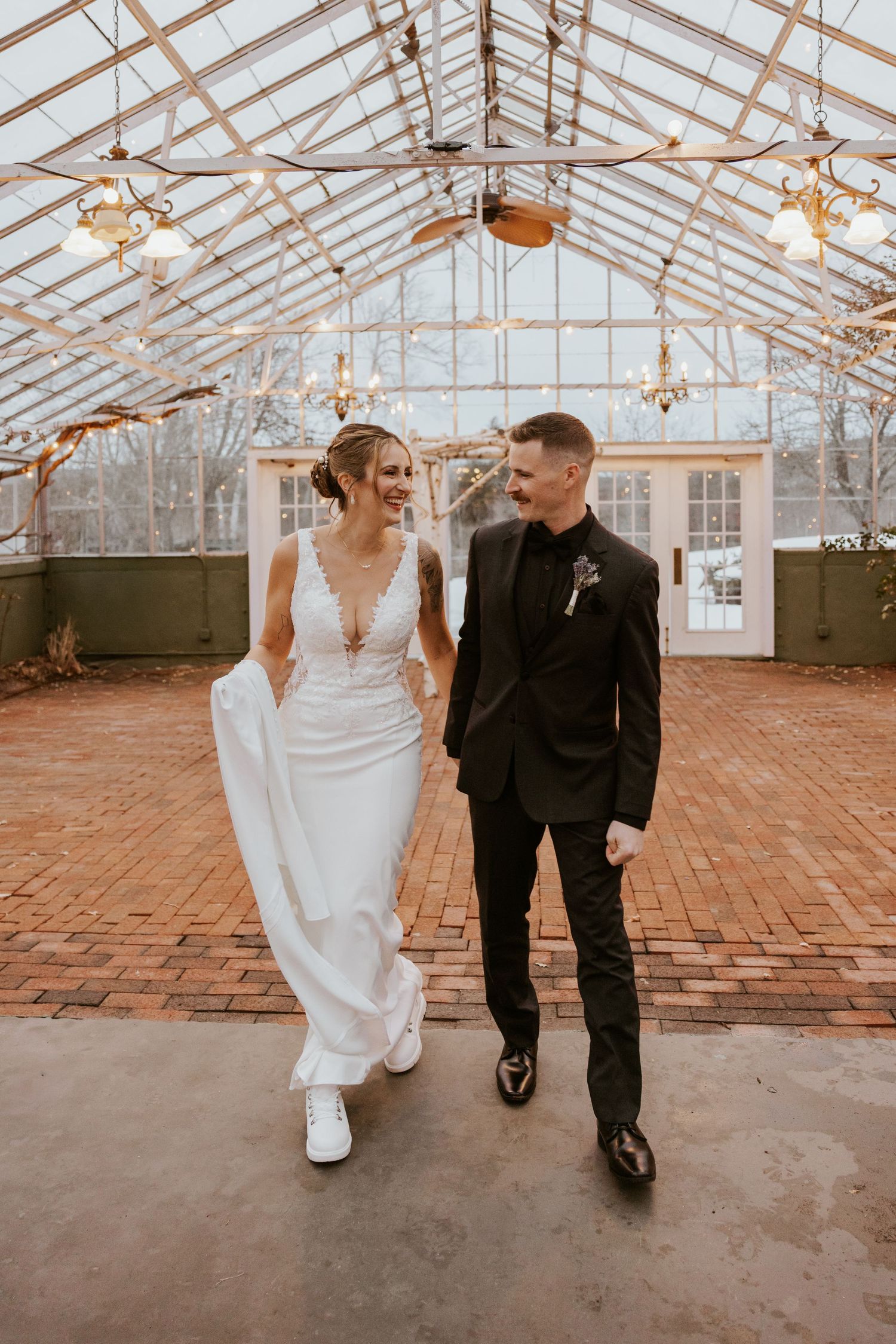 Bride and groom walking together in a glass greenhouse wedding venue, bride in white lace gown, groom in black suit.